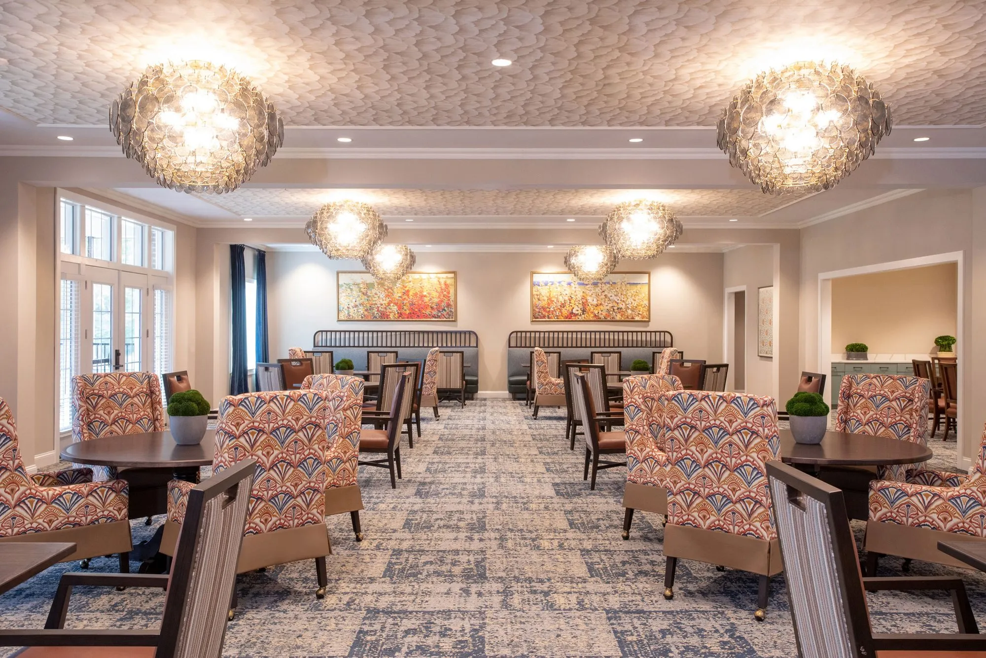 Senior living dining room under shell-like chandeliers, patterned armchairs at round tables and two banquettes