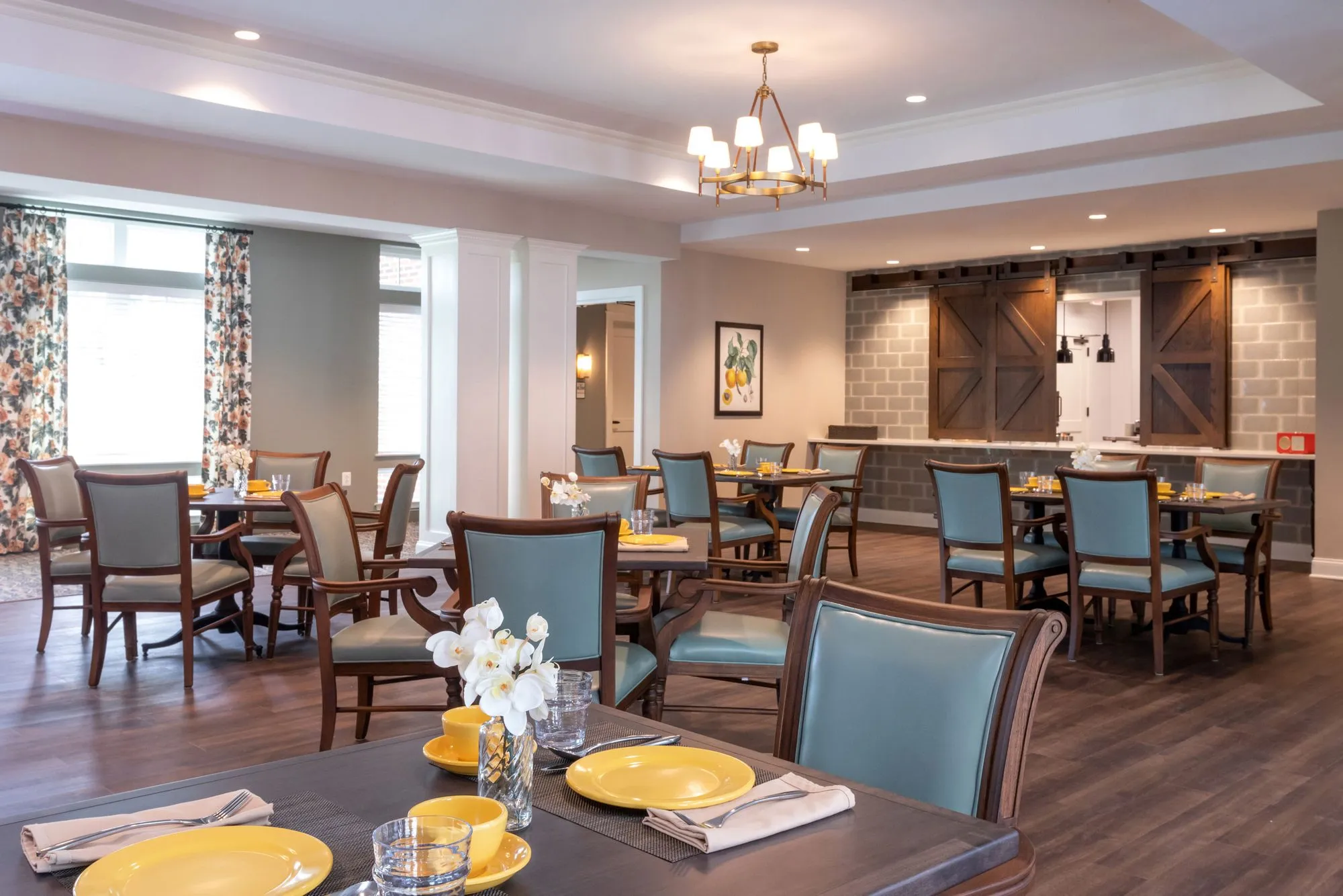 Senior living dining room with teal leather chairs at wood tables set with yellow plates, a brass chandelier and barn doors