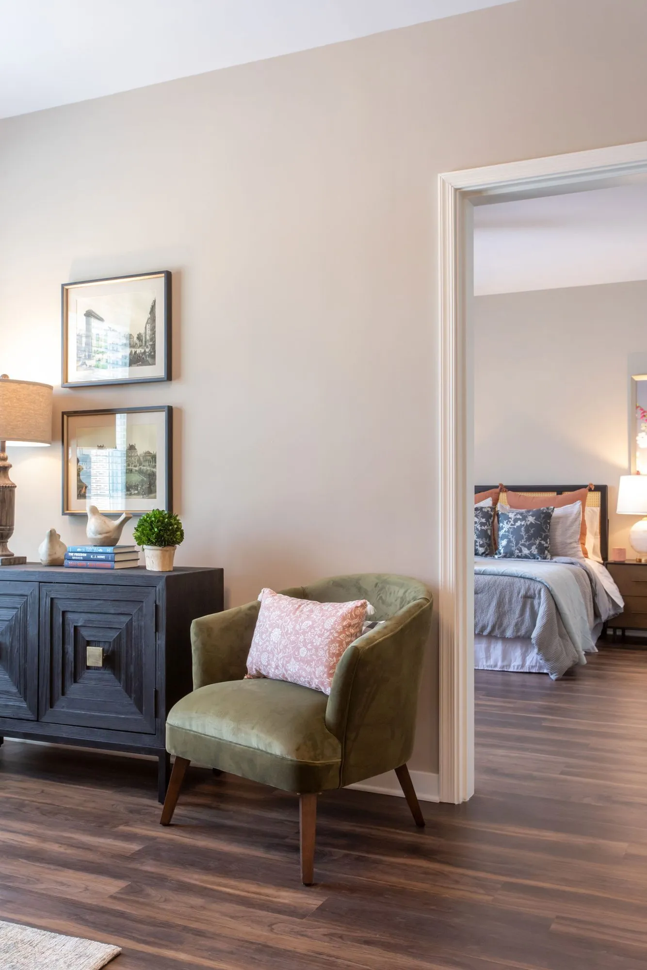 Senior living apartment entry with an olive velvet armchair beside a dark carved chest, the bedroom through the doorway