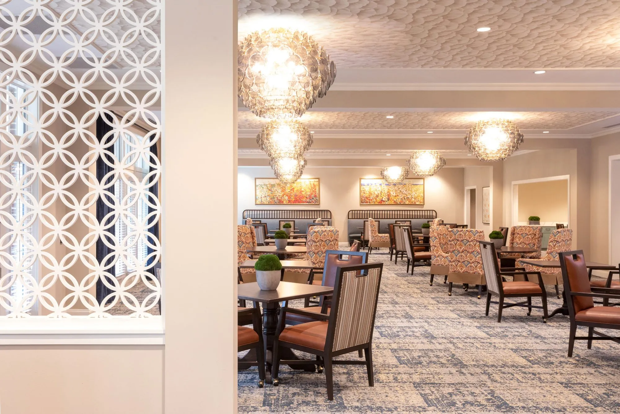 Senior living dining room seen past a white circle-pattern screen, shell chandeliers above striped and rust leather chairs