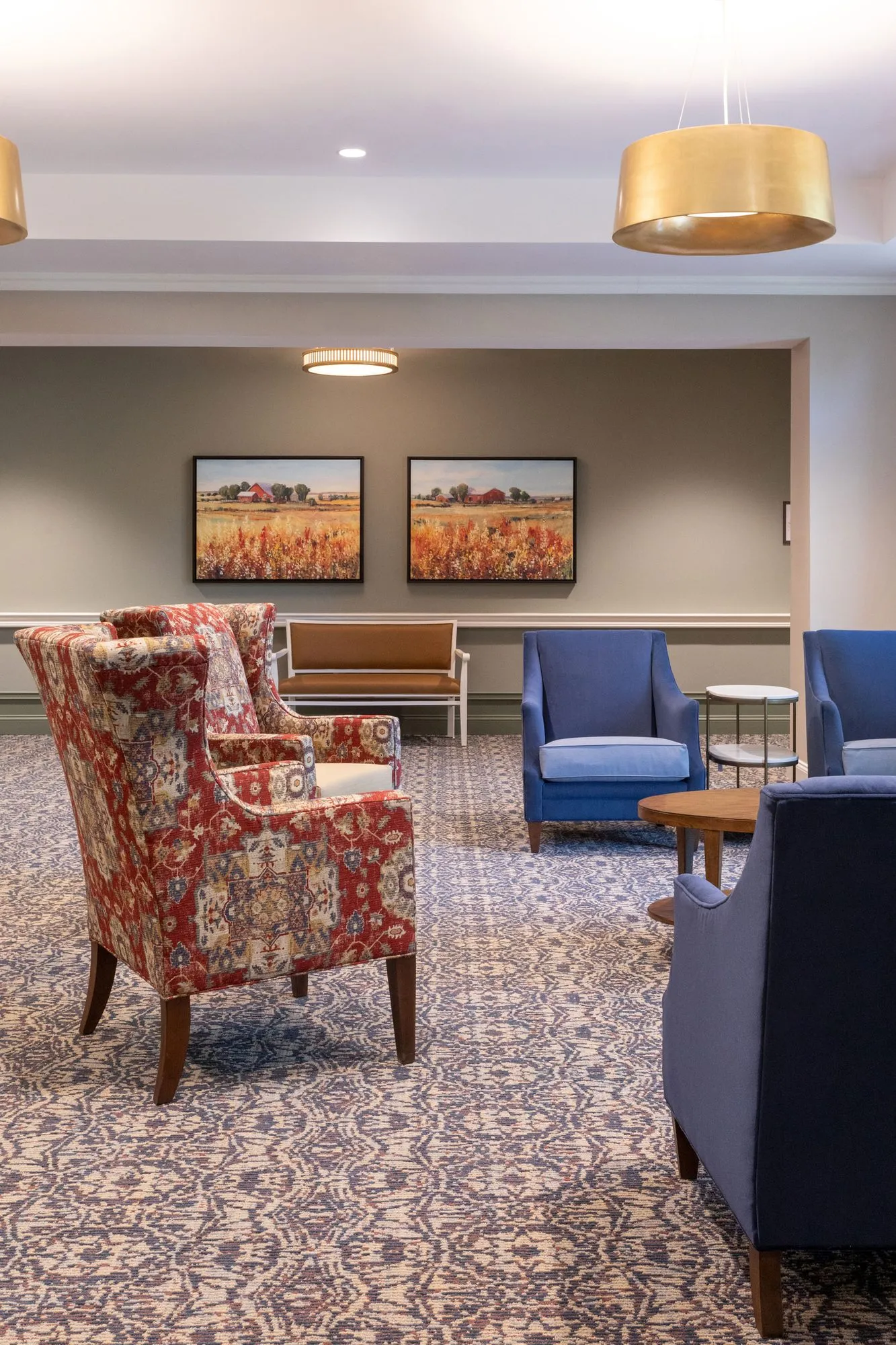 Senior living lounge with a red floral wing chair and blue armchairs on patterned carpet, field paintings and gold pendants