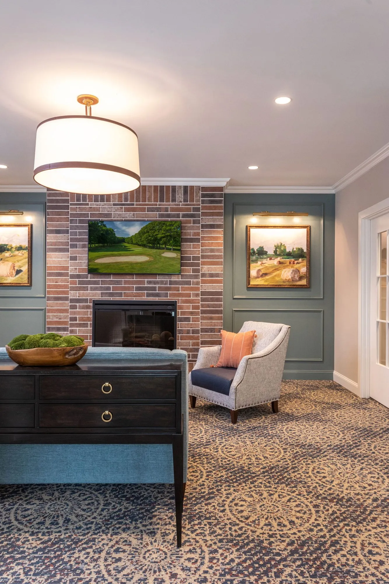 Senior living lounge with a brick fireplace and golf course painting between sage panelled walls under a drum pendant