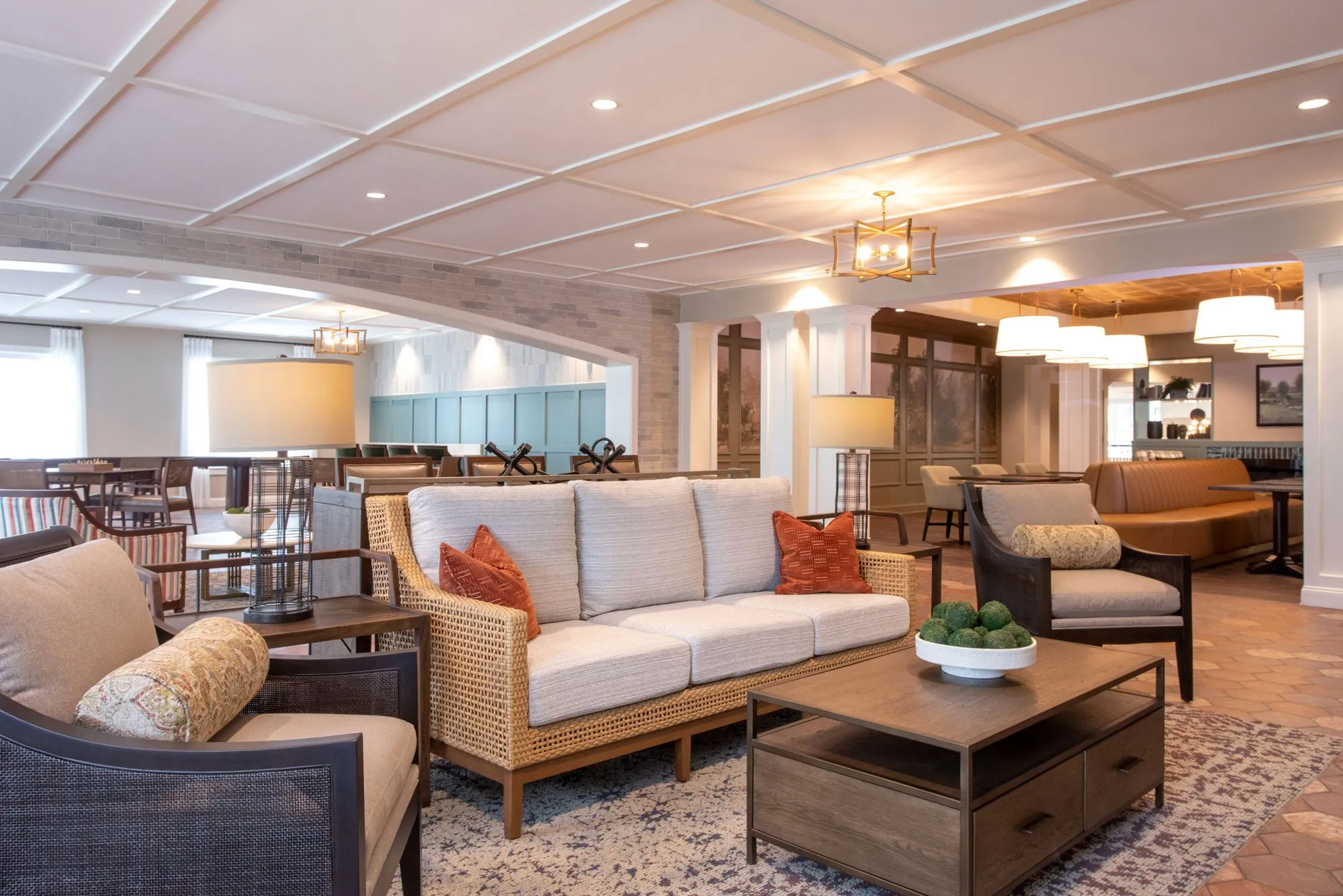 Senior living lounge with a woven-frame sofa and cane chairs on terracotta tile, the pendant-lit bistro beyond