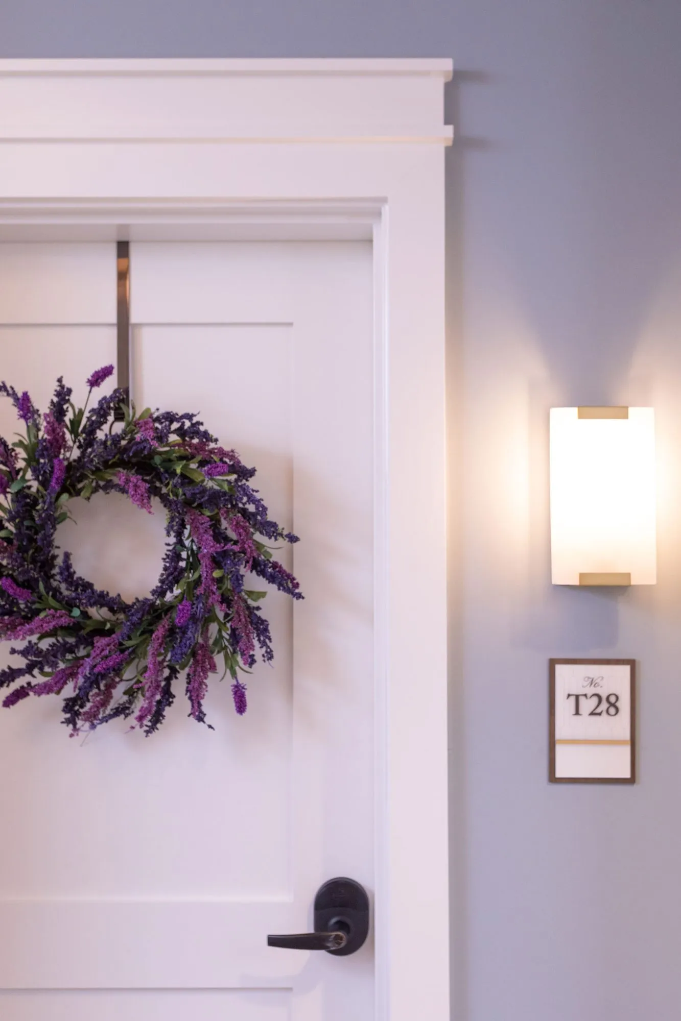 Senior living apartment door with a lavender wreath beside a wall sconce and a T28 number plaque