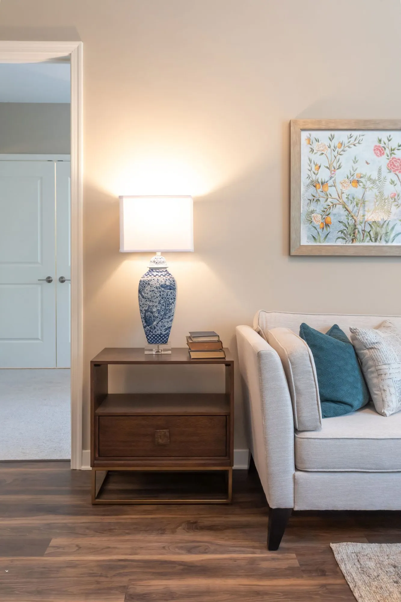 Senior living living room corner with a linen sofa, teal pillows and a blue and white lamp below a floral print