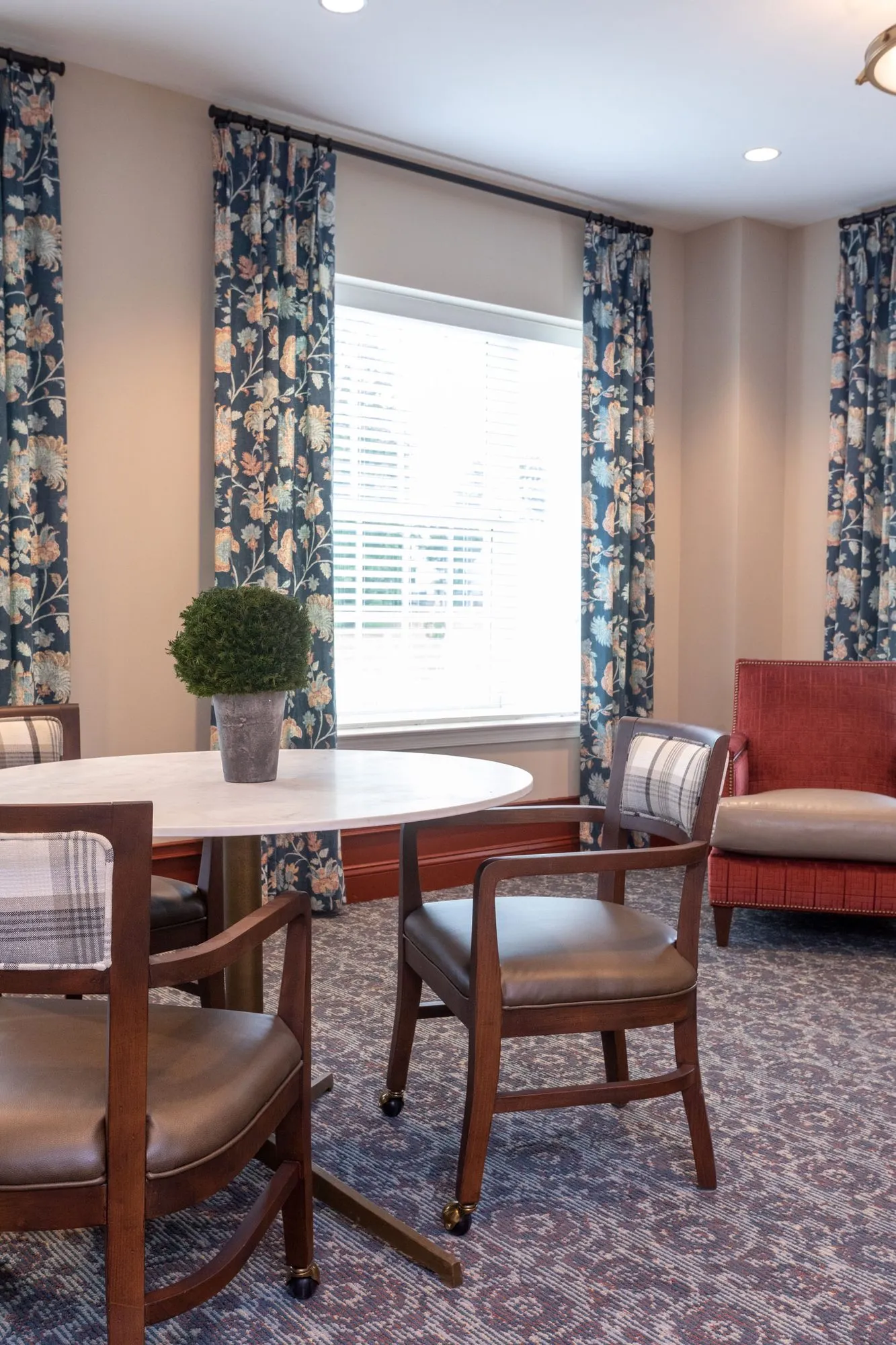 Senior living interior detail: round marble table and leather chairs before windows with navy floral drapery, a red armchair