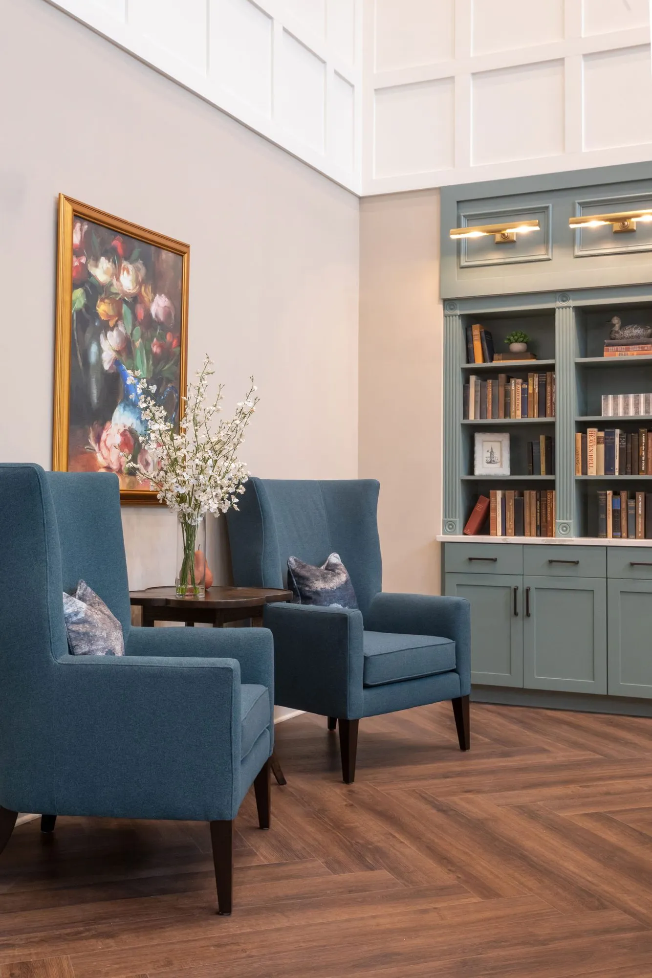 Senior living interior detail: two blue wing chairs beside a floral still life and sage bookcases with picture lights