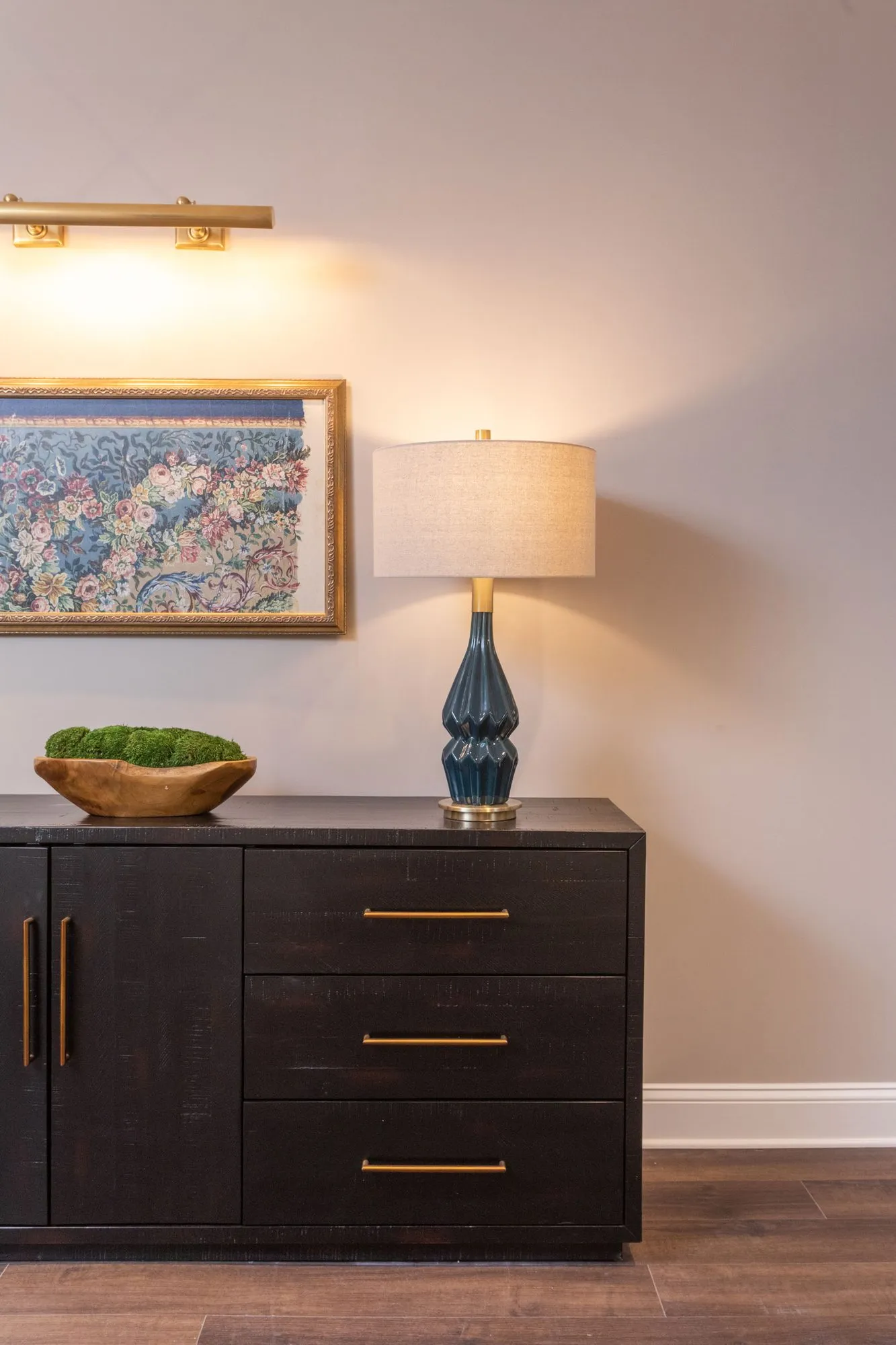 Senior living interior detail: black sideboard with a navy lamp and moss bowl under a floral painting with a picture light