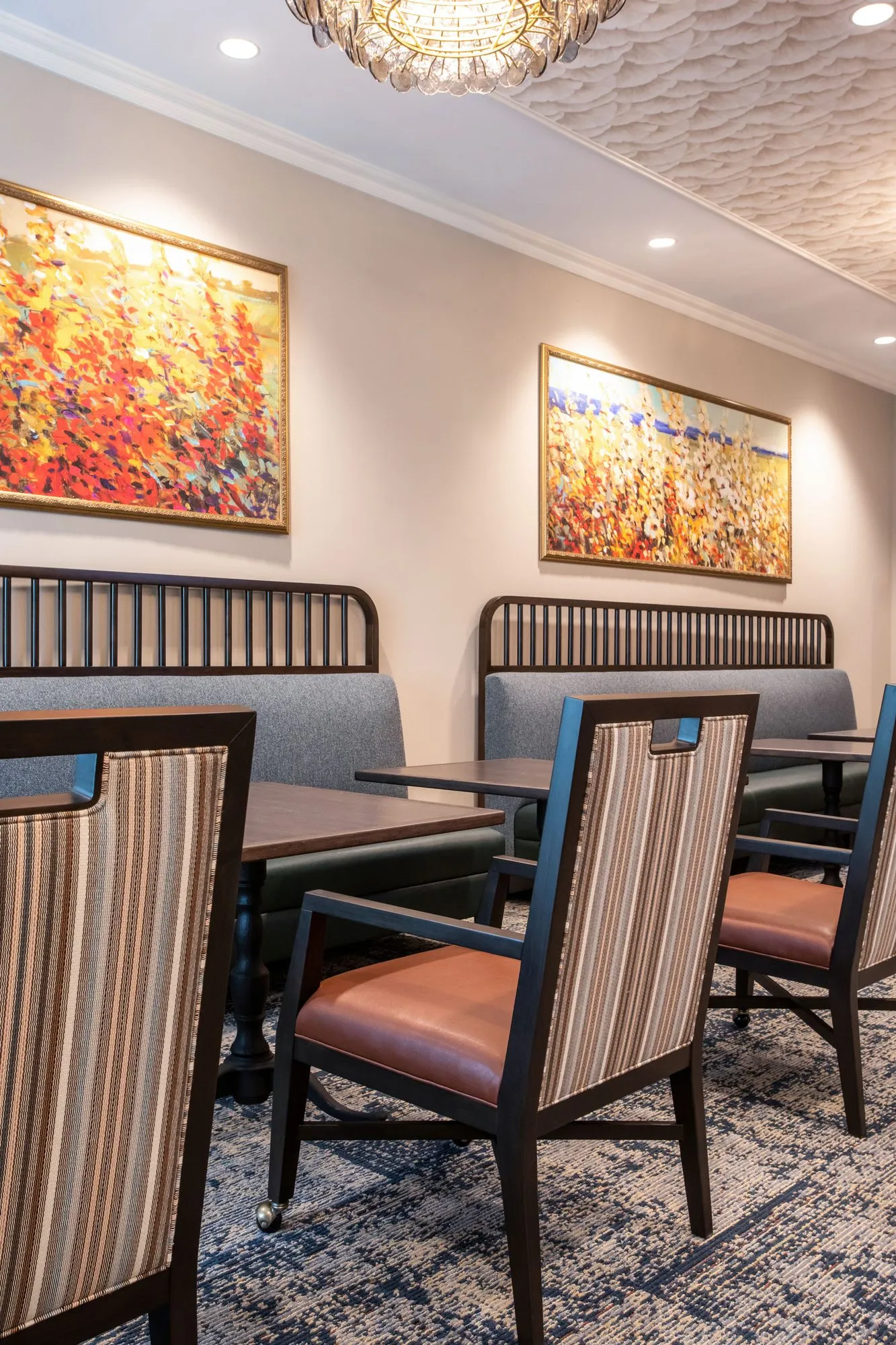 Senior living dining room with spindle-back banquettes below floral landscape paintings, striped chairs at dark tables