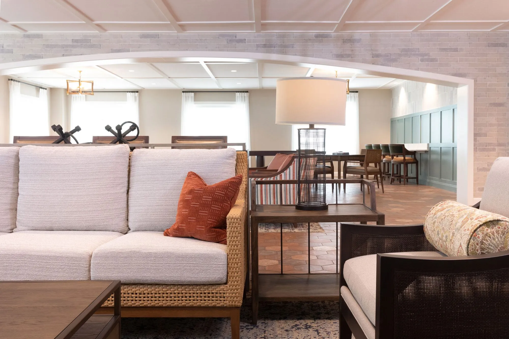 Senior living lounge seen past a linen sofa with a rust pillow toward an arched brick opening and the dining room beyond