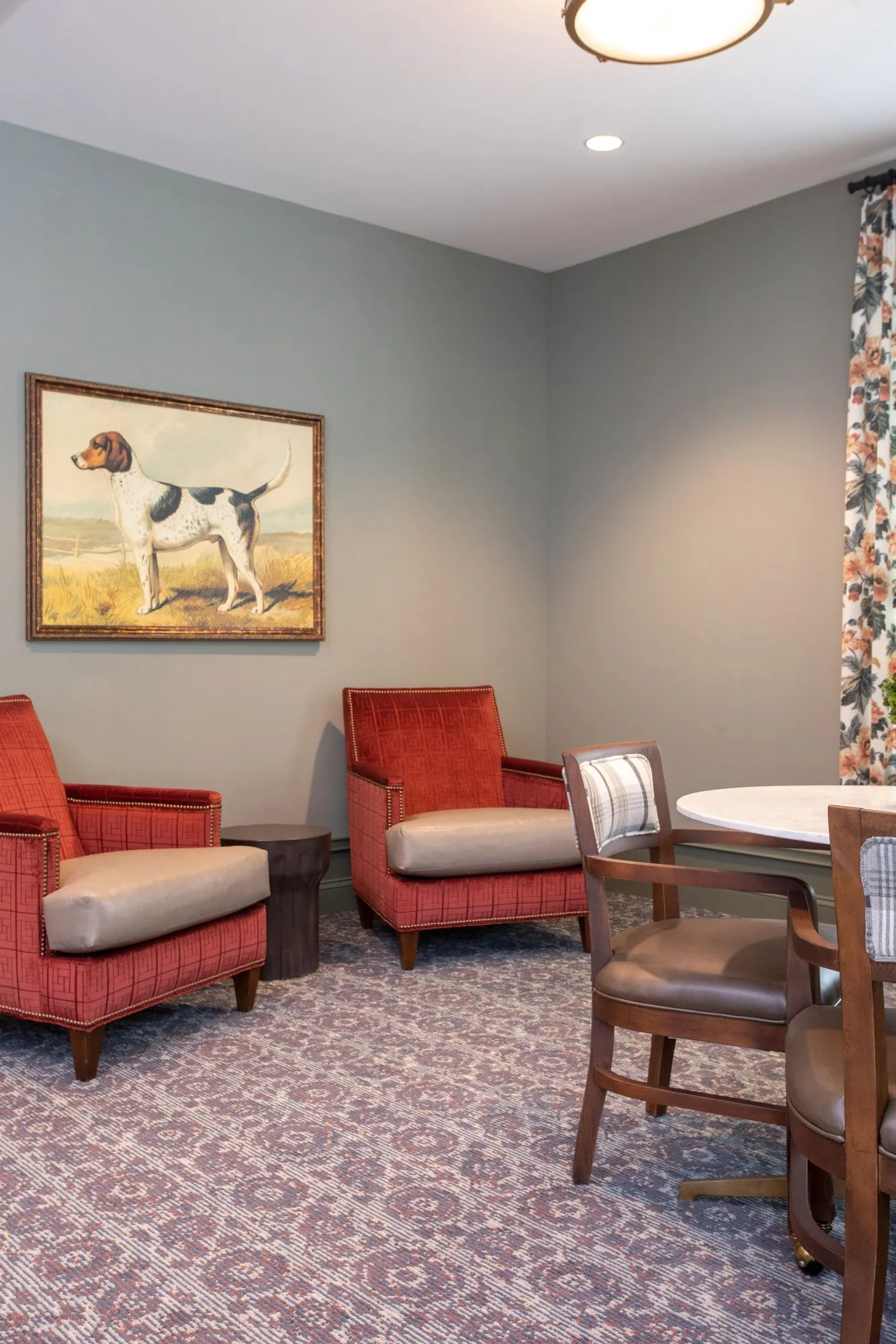 Senior living card room with red armchairs and leather chairs at a round table below a painting of a hound
