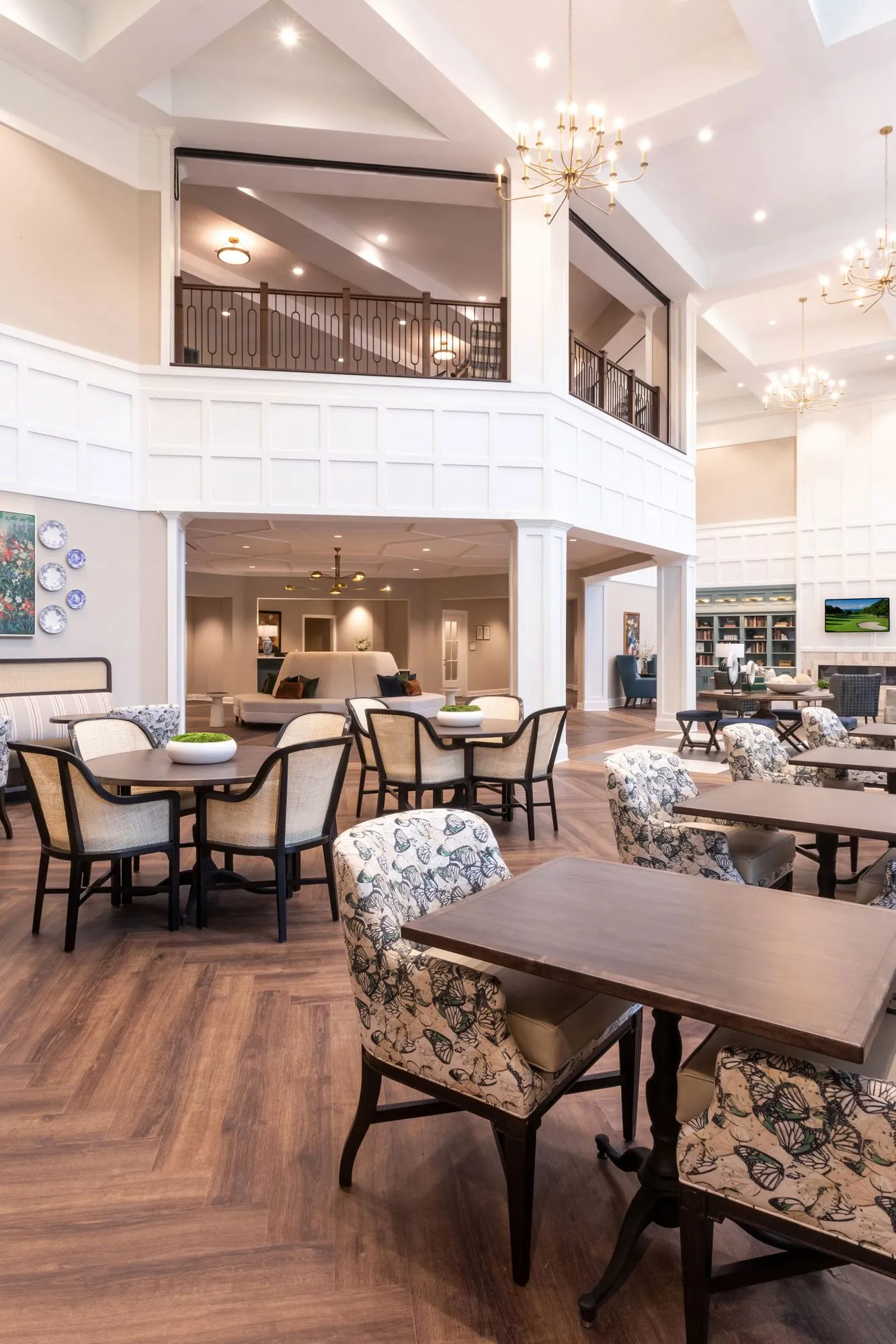 Senior living double-height dining room with floral armchairs at wood tables beneath the railed balcony