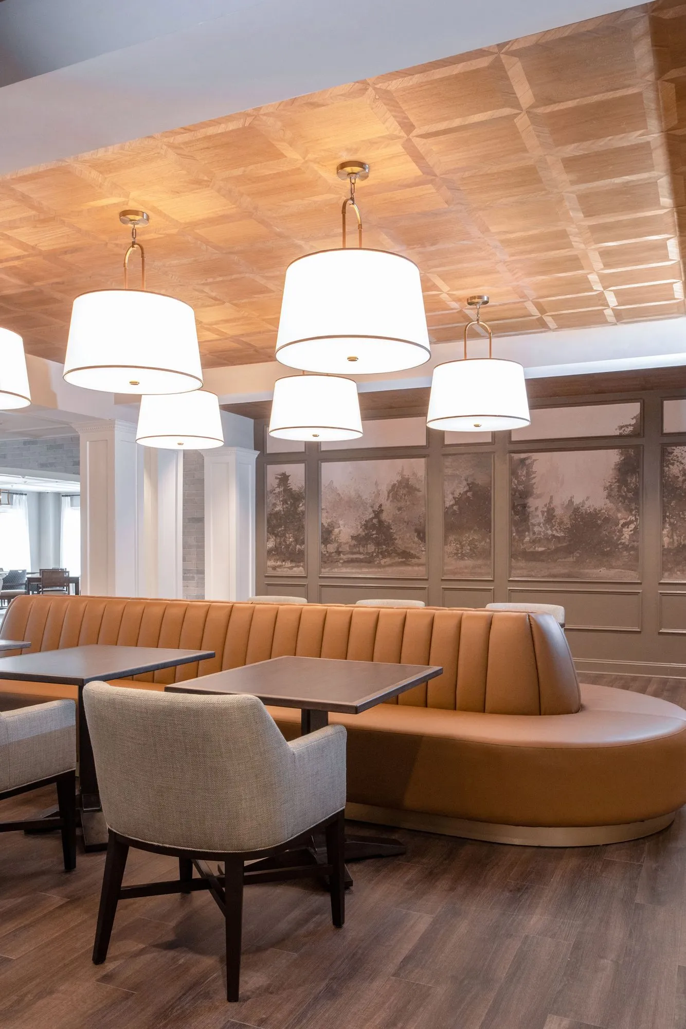 Senior living bistro with a curved cognac banquette and tables under white drum pendants and a coffered wood ceiling