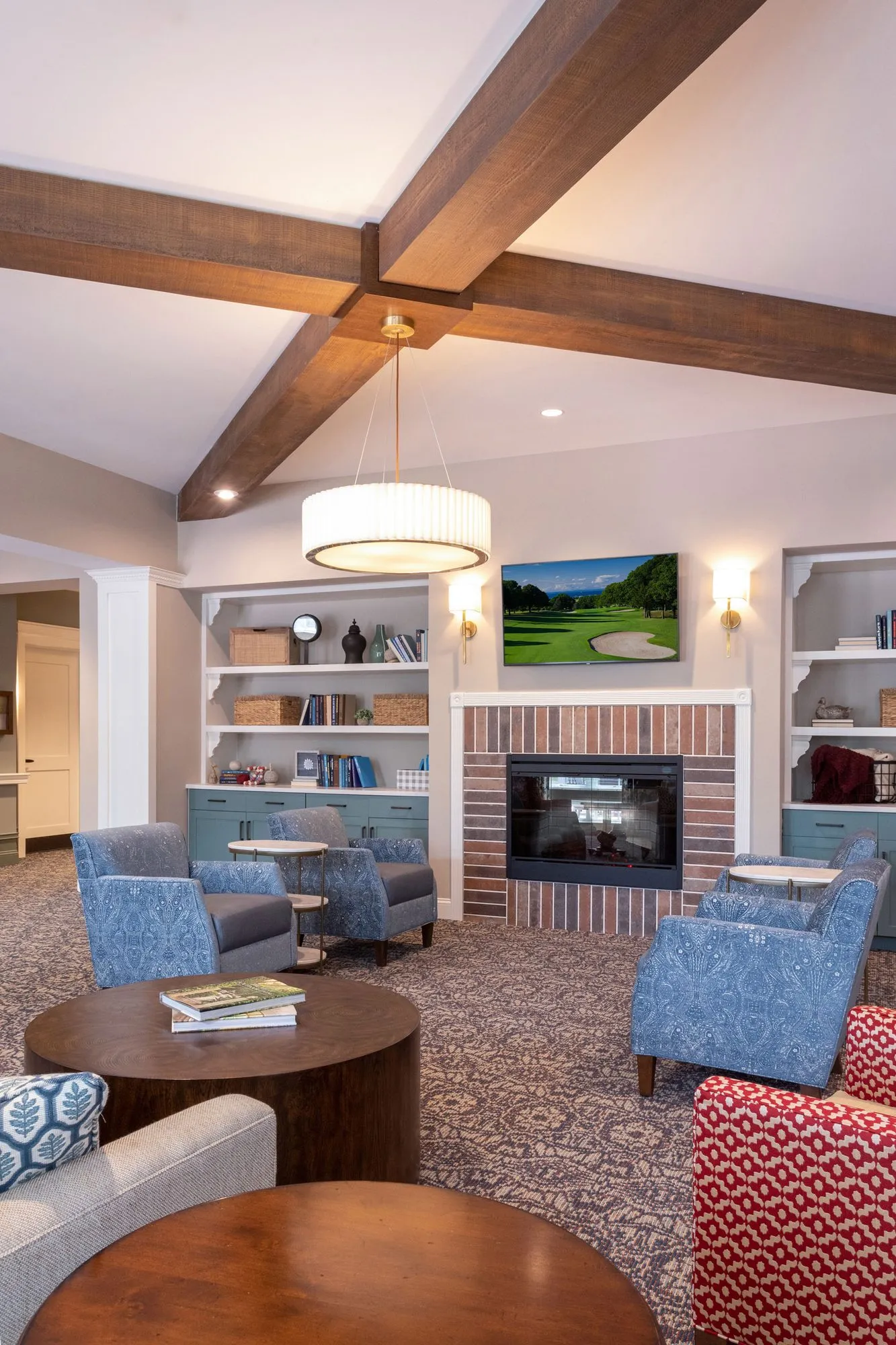 Senior living lounge under timber beams with blue armchairs around a brick fireplace, a golf course painting and bookcases