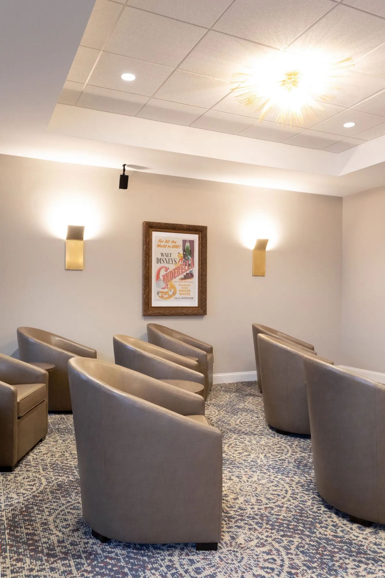 Senior living theater with rows of taupe leather barrel chairs facing a framed vintage film poster between brass sconces