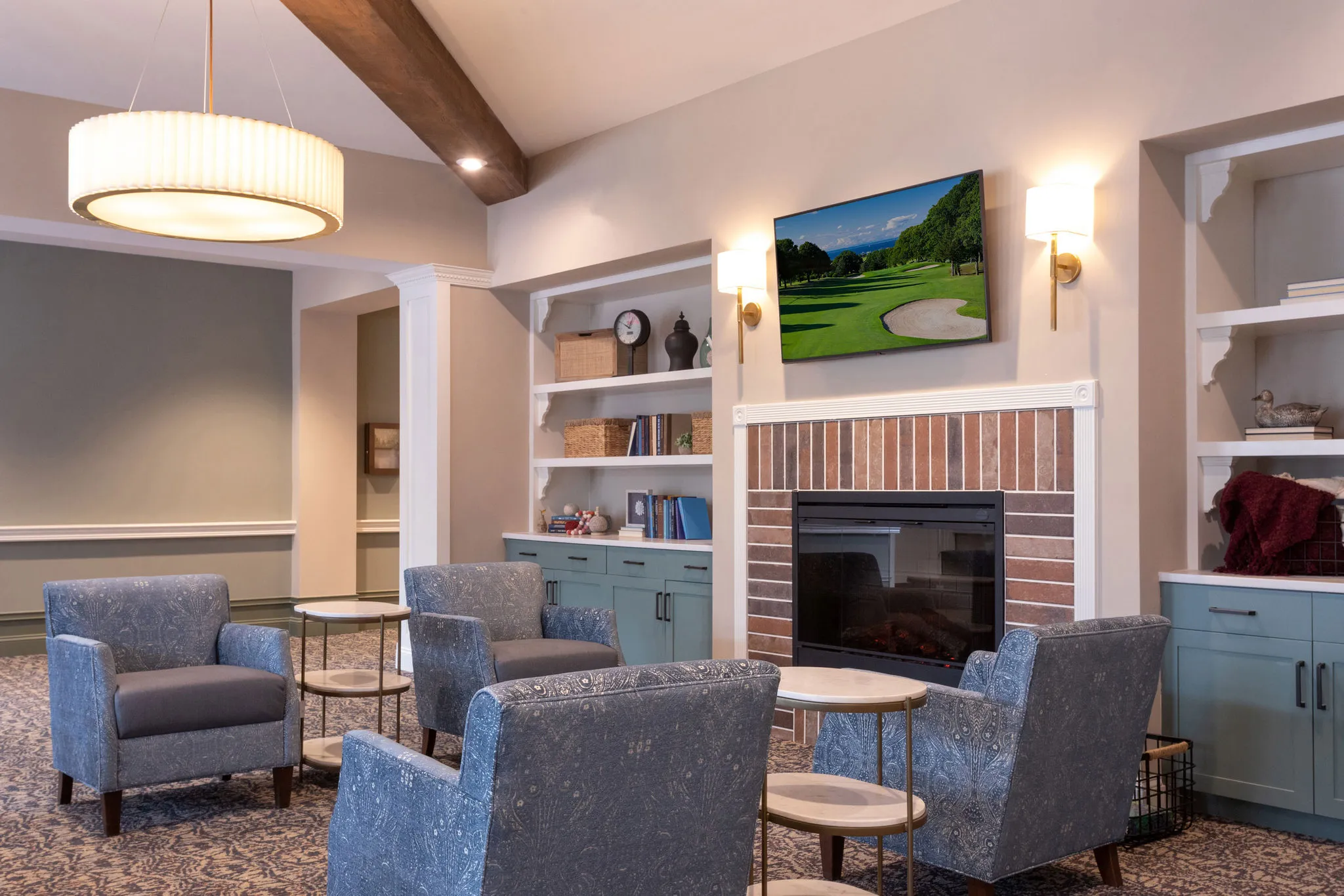 Senior living club room with a brick fireplace flanked by sage cabinetry and blue armchairs around drink tables