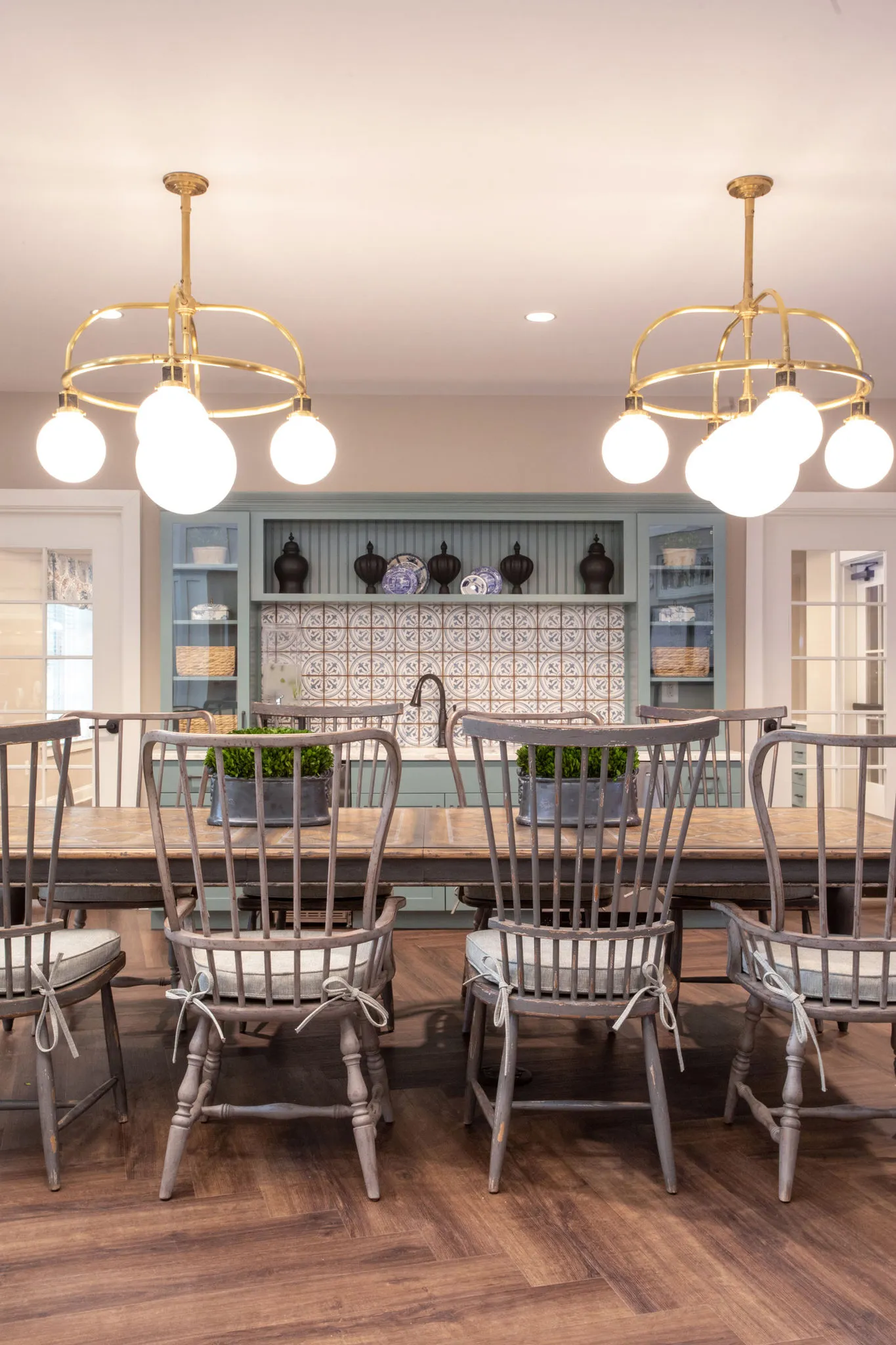 Senior living country kitchen with spindle-back chairs at a long wood table below brass globe chandeliers, patterned tile