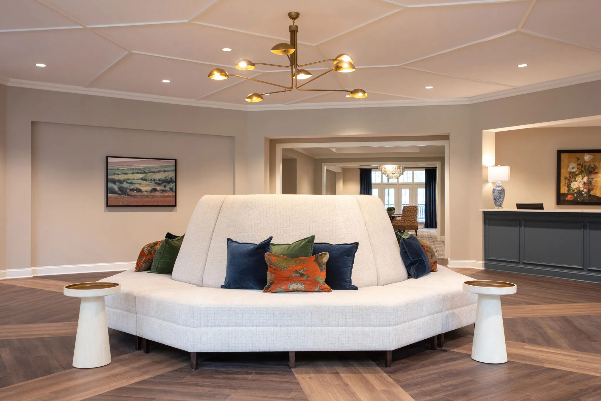 Senior living lobby with a curved white settee below a brass multi-arm chandelier, the reception desk at right
