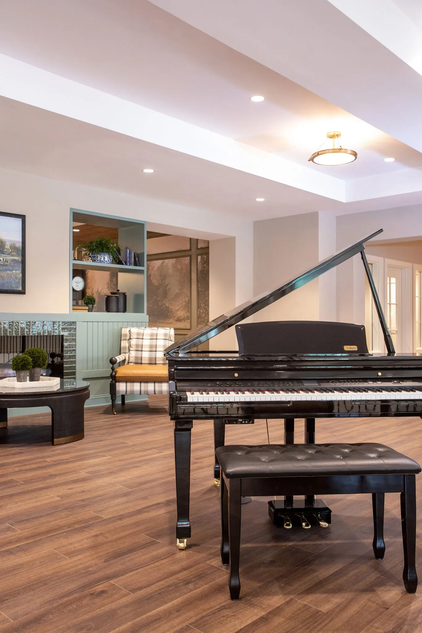 Senior living parlor with a grand piano on the wood floor, a plaid armchair by a tiled fireplace beyond