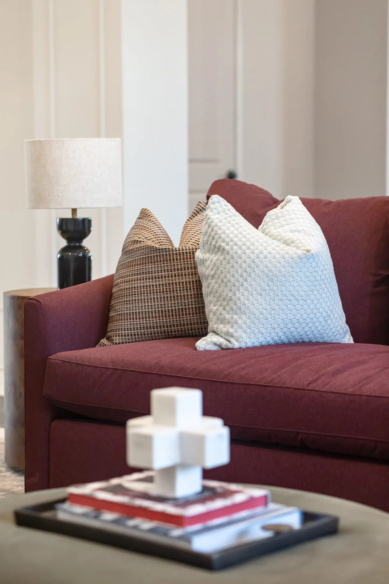 Multifamily amenity interior detail: white sculptural cross on stacked books on an ottoman tray, a burgundy sofa behind