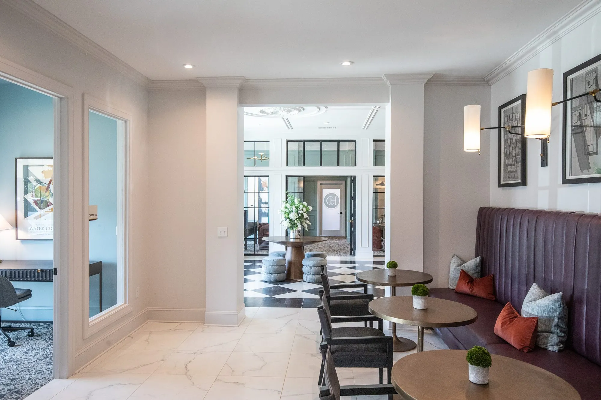 Multifamily amenity corridor with a plum channel-tufted banquette and cafe tables, a glass-walled office at left