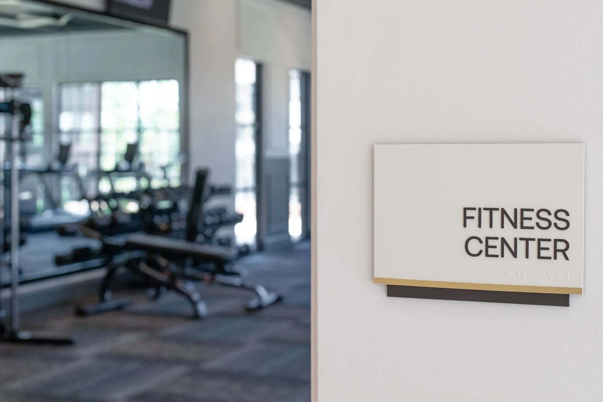 Multifamily amenity fitness center entry with a wall sign reading Fitness Center by the glass, weight benches inside