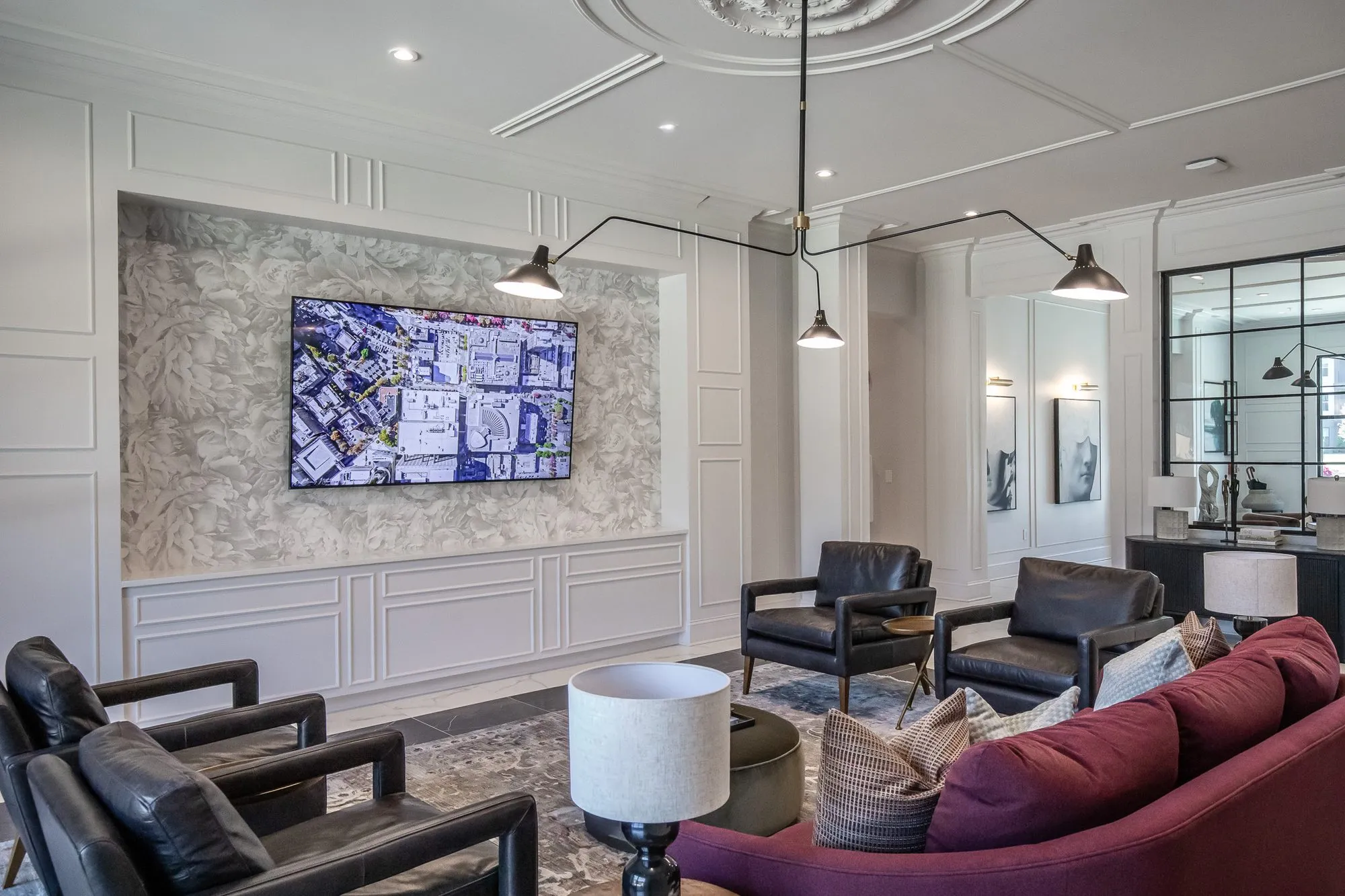 Multifamily amenity clubroom with a burgundy sofa and black leather chairs facing a wall screen in floral relief wallpaper