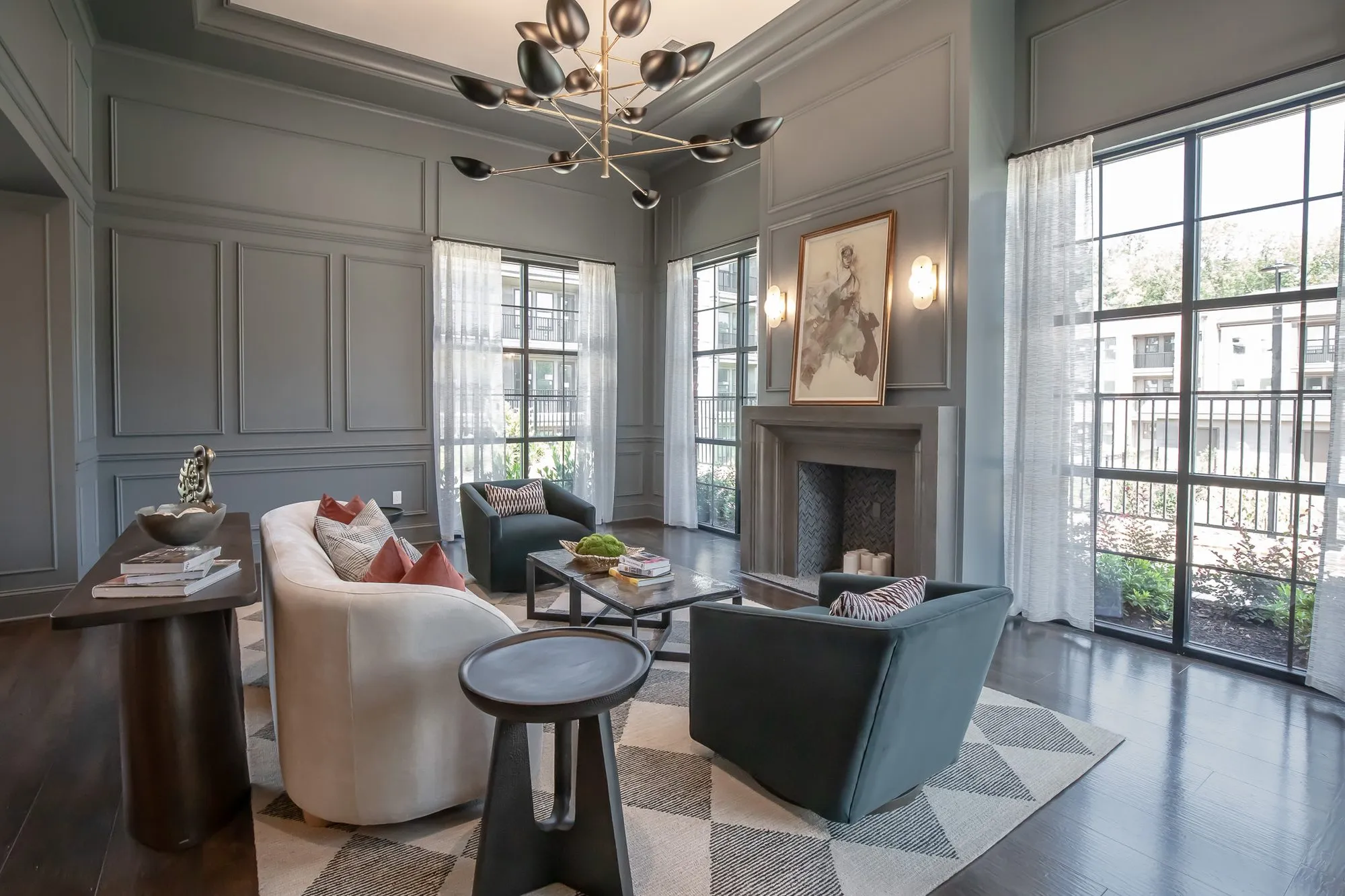 Multifamily amenity sitting room with a white curved sofa and teal chairs before a fireplace under a brass bird chandelier