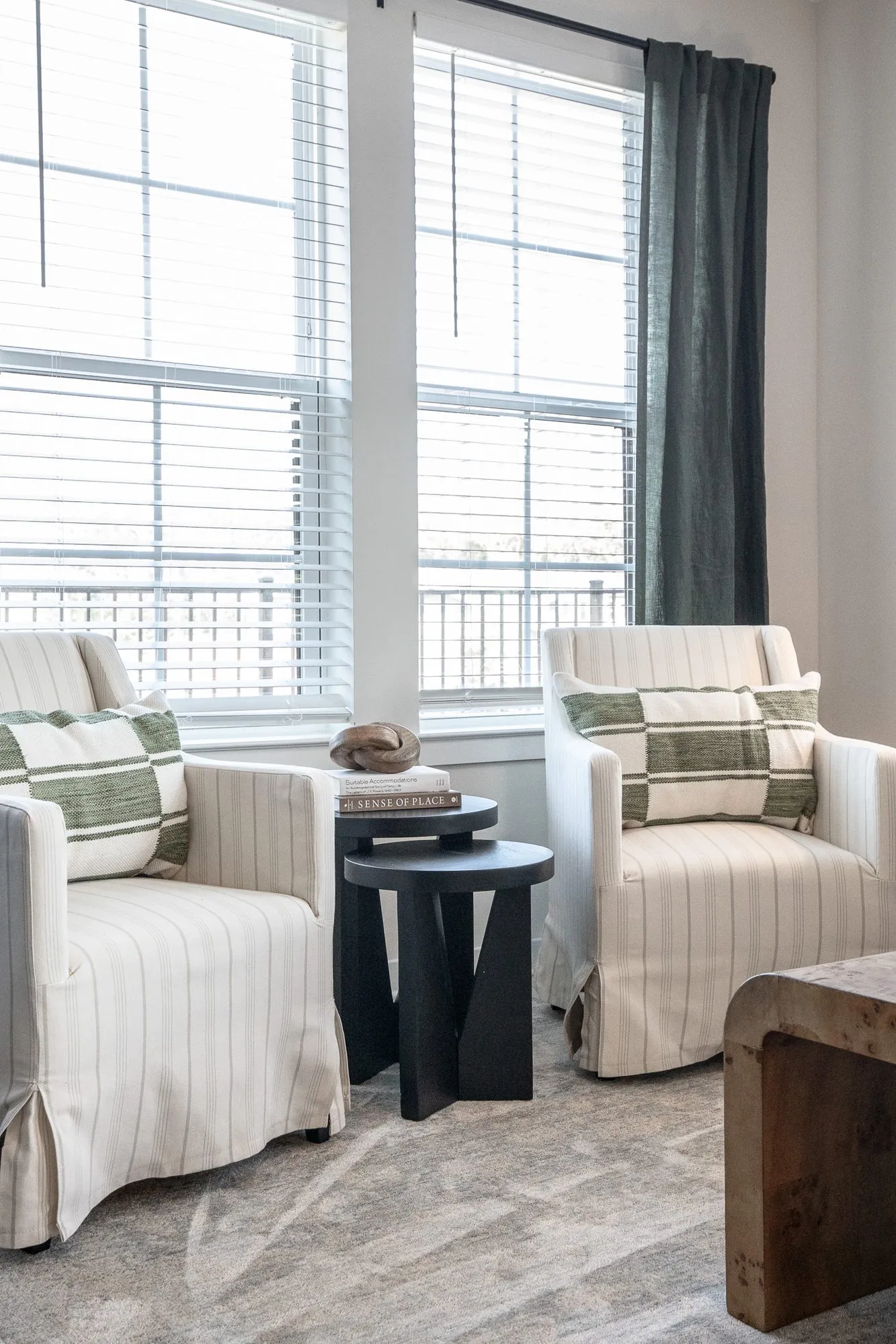 Multifamily interior detail: two striped linen slipcovered armchairs with green pillows by a window, a black side table