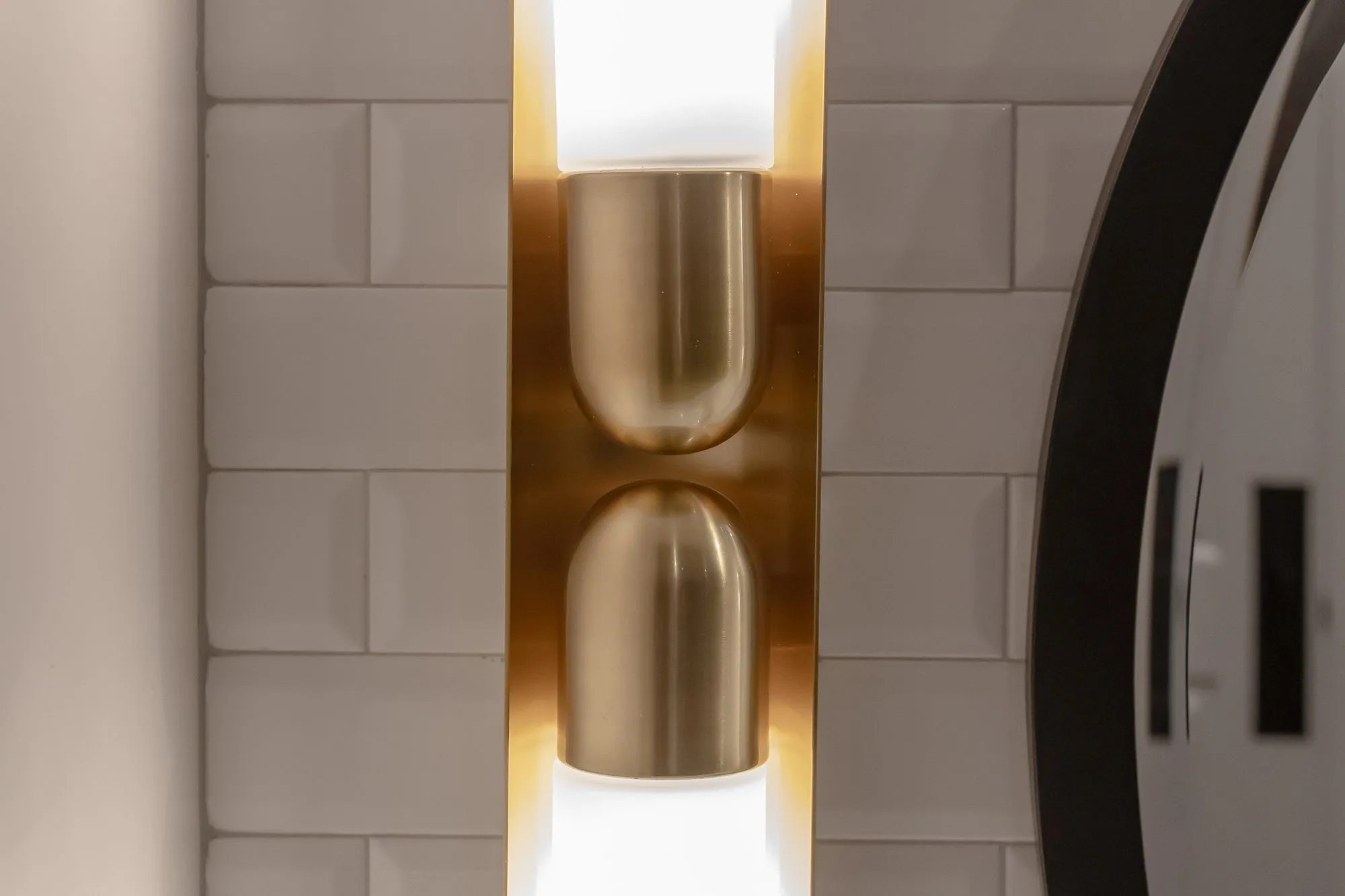 Multifamily interior detail: brass up-and-down wall sconce on white dimensional tile beside the edge of a round mirror