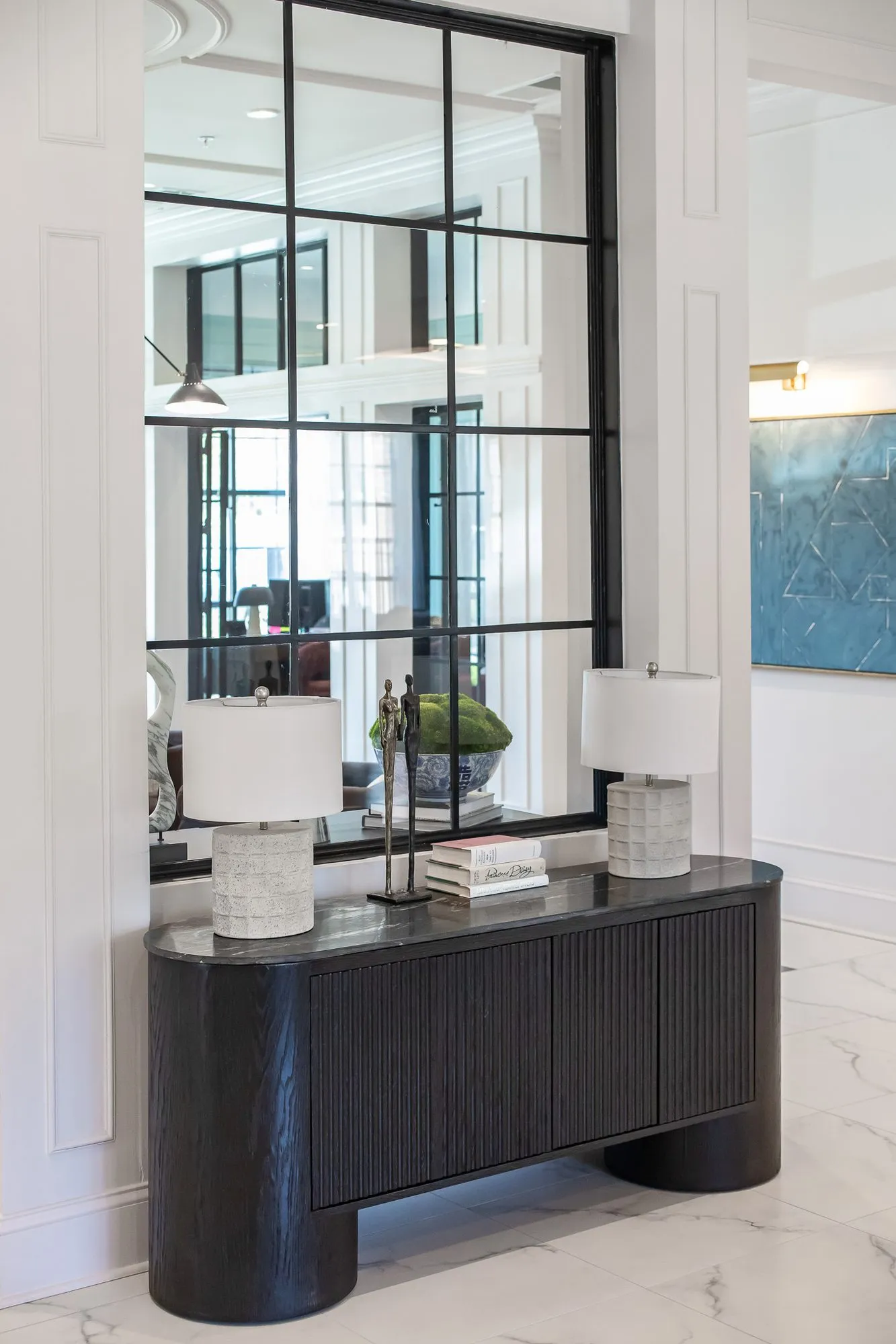 Multifamily amenity lobby lounge with a black fluted console and two white lamps below a black grid window mirror