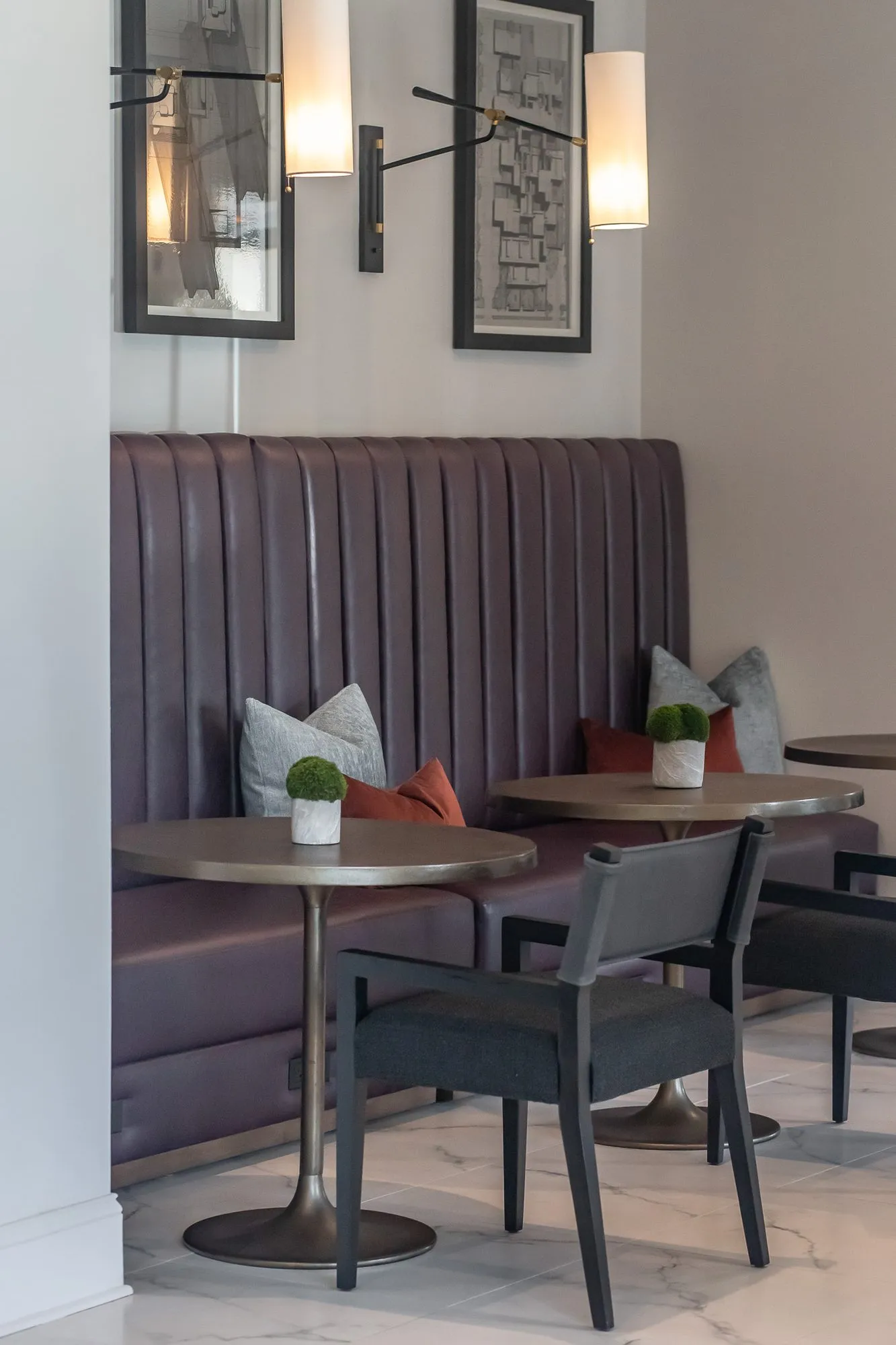 Multifamily amenity corridor with a plum channel-tufted banquette, cafe tables and black chairs below framed prints