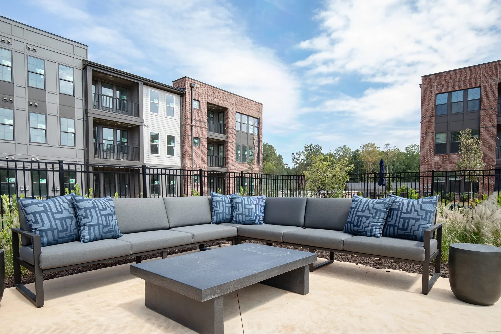Multifamily community outdoor lounge with a sectional and blue patterned pillows around a concrete table, apartments beyond
