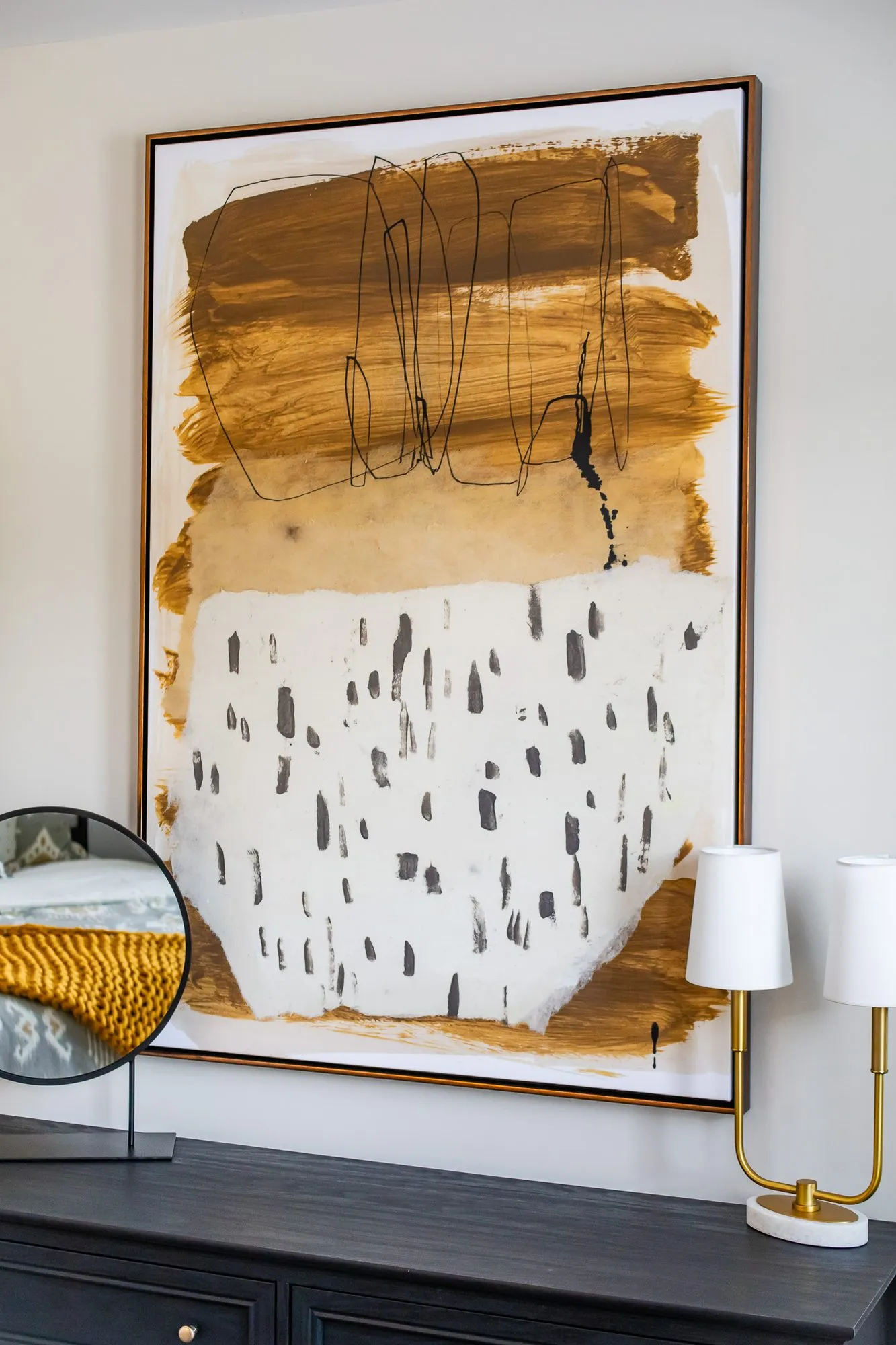 Multifamily interior detail: large abstract painting in ochre and black brushstrokes above a black dresser with a brass lamp