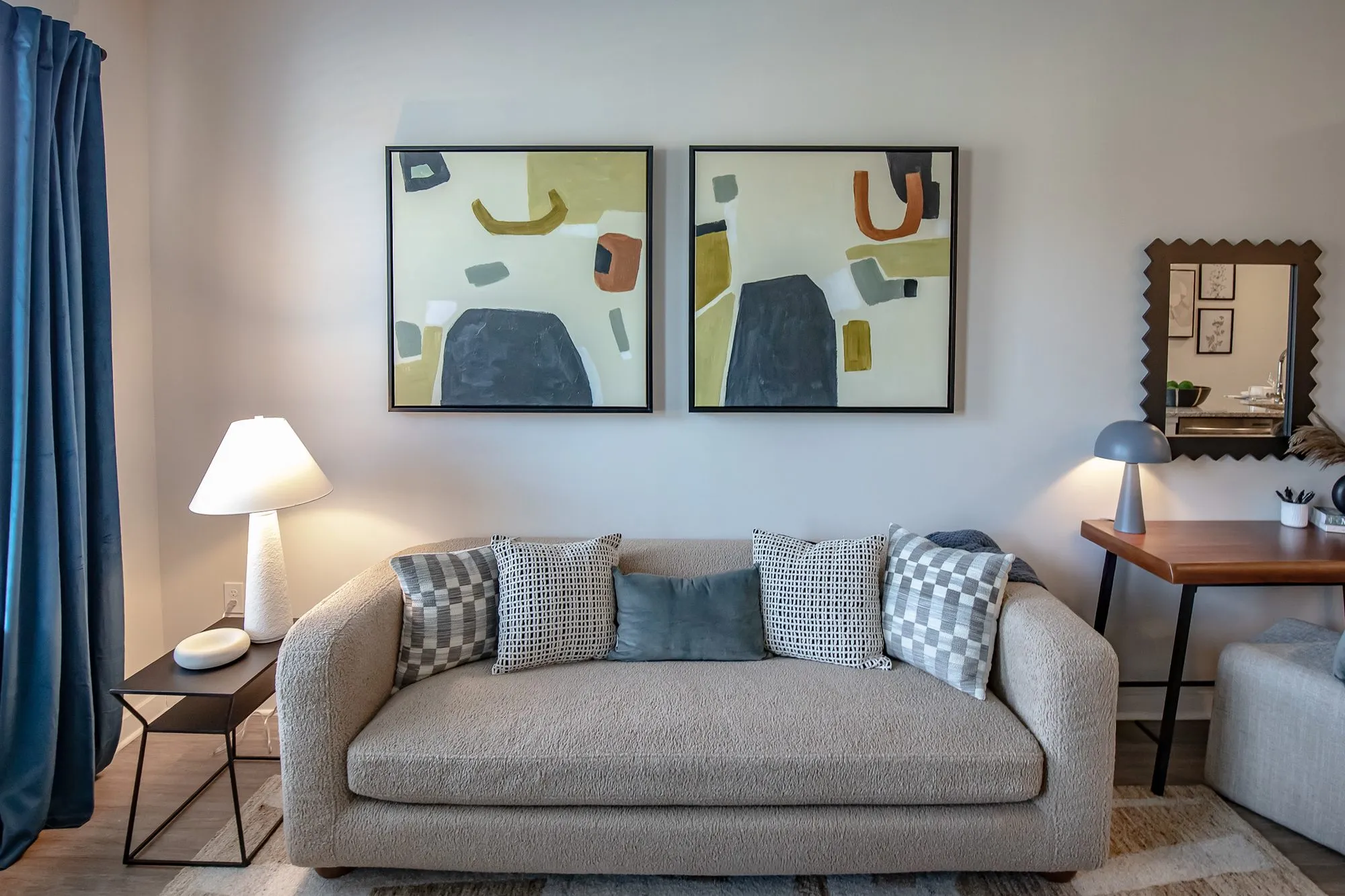 Multifamily apartment model living room with a taupe sofa and patterned pillows below two abstract prints, a blue curtain