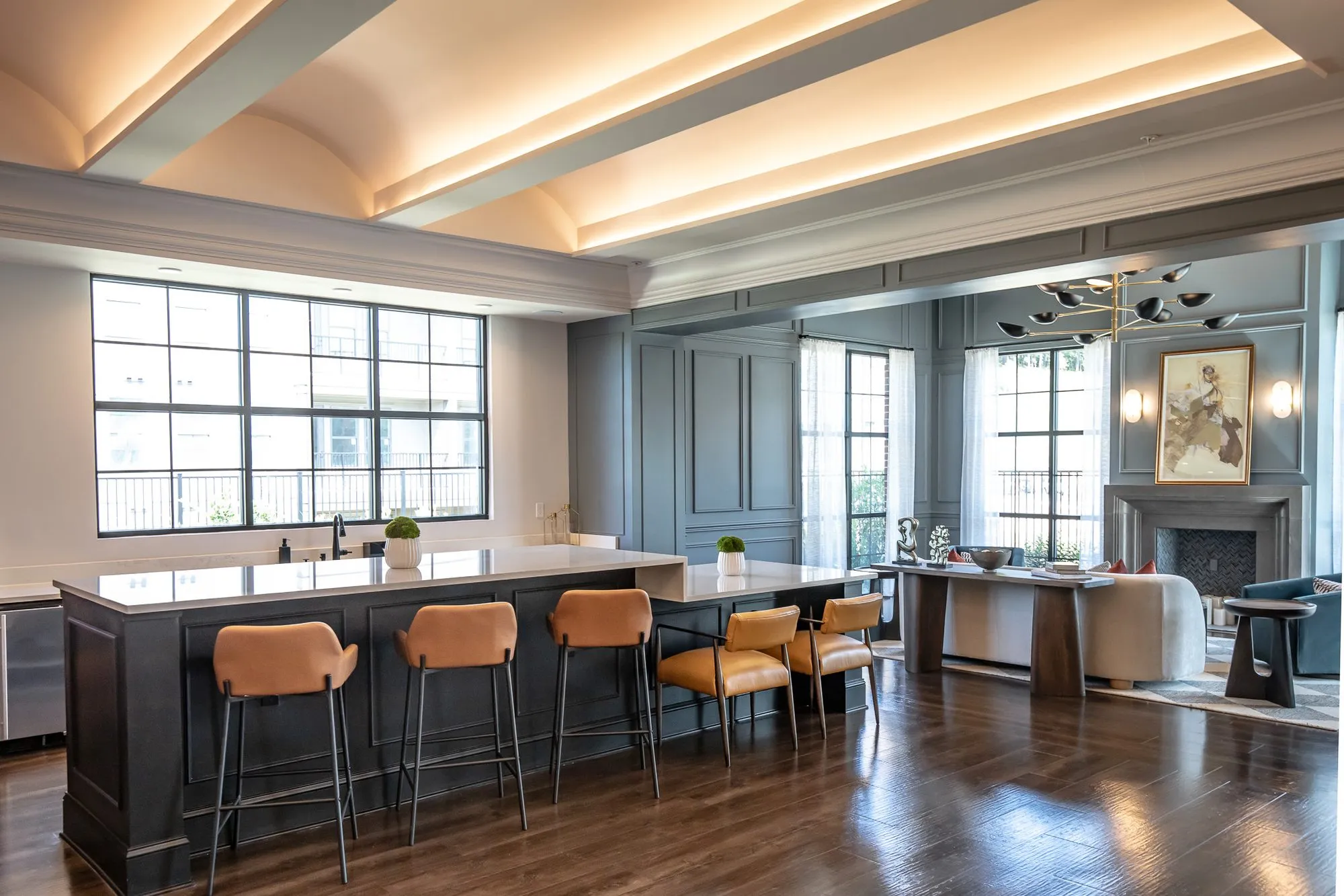 Multifamily amenity clubroom with a long dark kitchen island and cognac leather stools under a vaulted tray ceiling