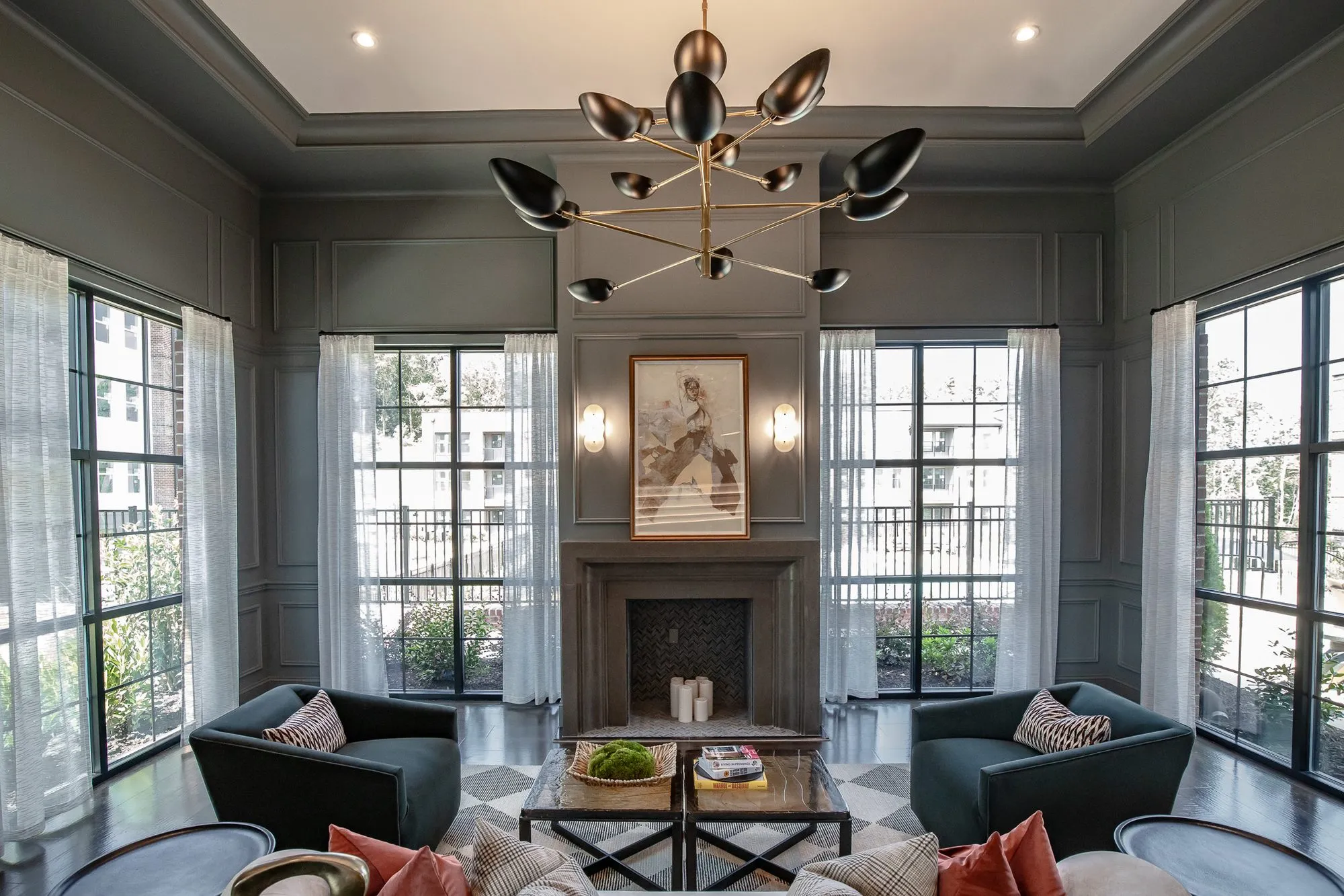 Multifamily amenity sitting room with teal chairs facing a fireplace between tall windows under a brass bird chandelier