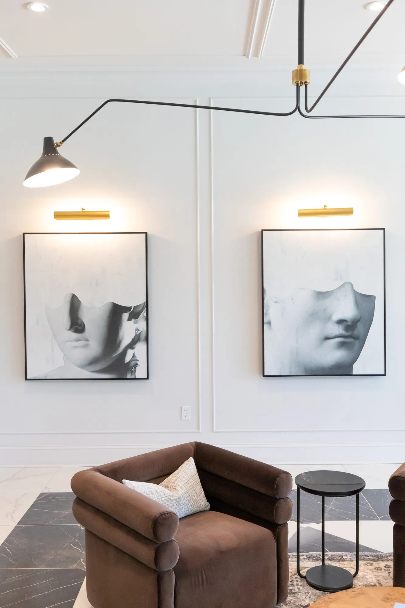 Multifamily amenity lobby lounge with a brown velvet barrel chair below two classical face prints lit by brass picture lights