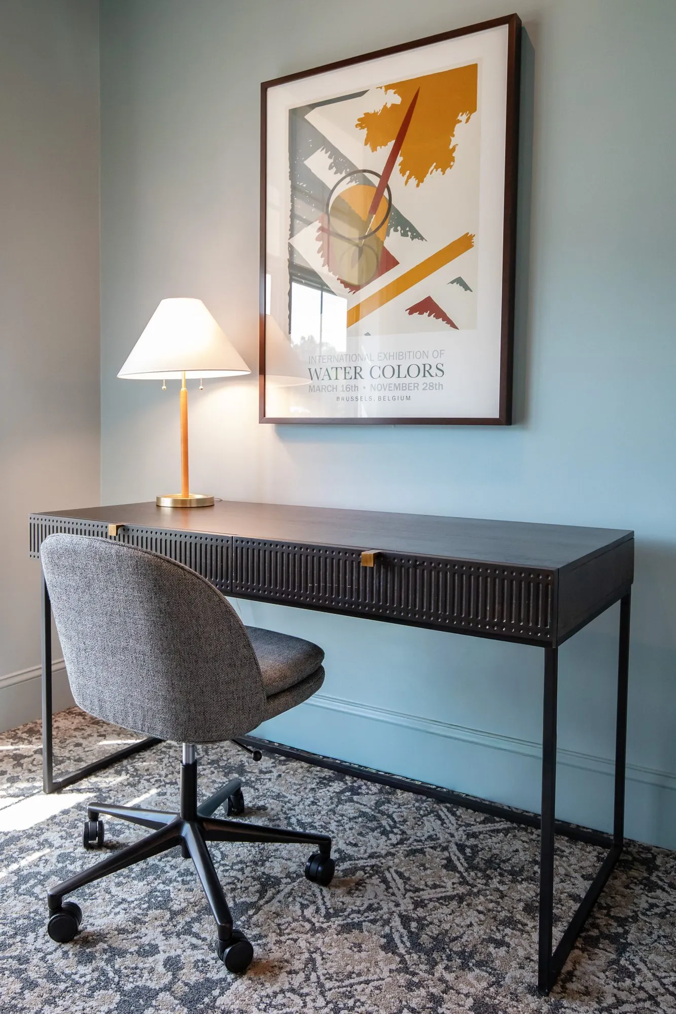 Multifamily interior detail: desk with a grey task chair and brass lamp below a watercolour exhibition poster on a blue wall