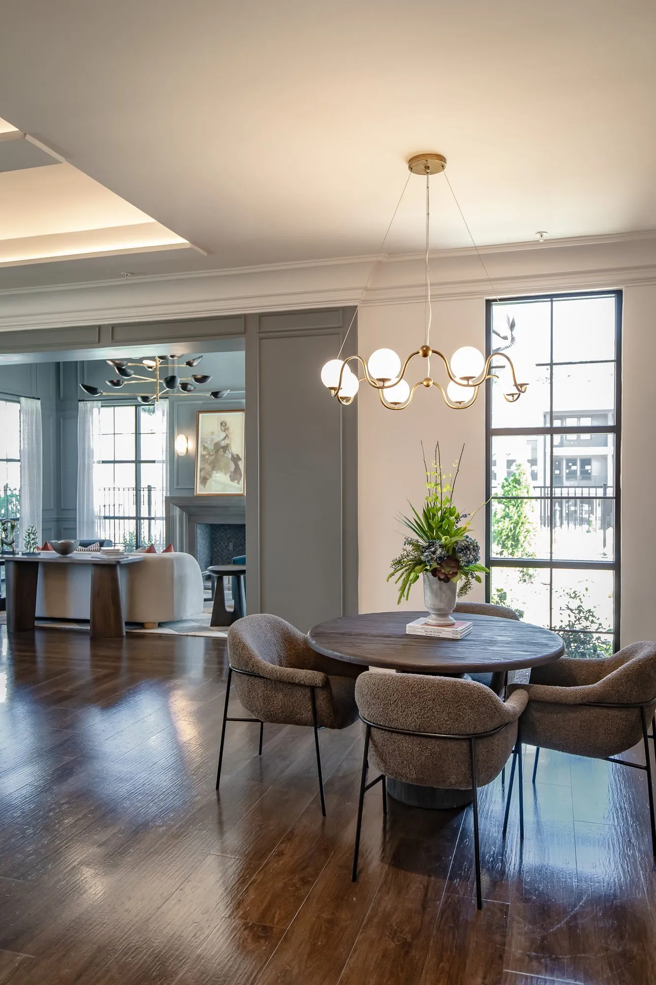 Multifamily amenity dining area with grey woven chairs at a round table under a brass globe chandelier by a tall window