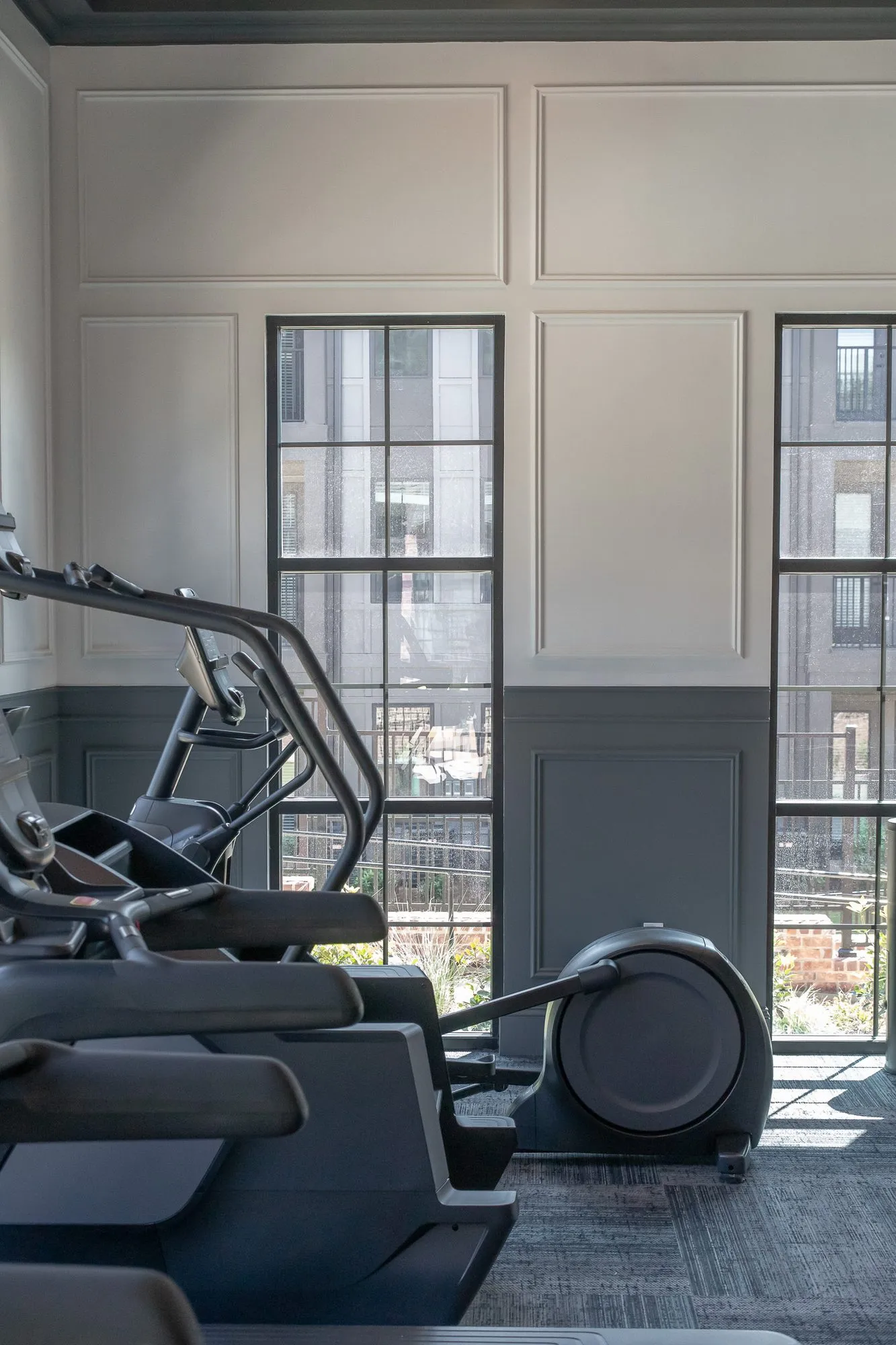 Multifamily amenity fitness room with cardio machines facing tall black framed windows, white and grey panelling
