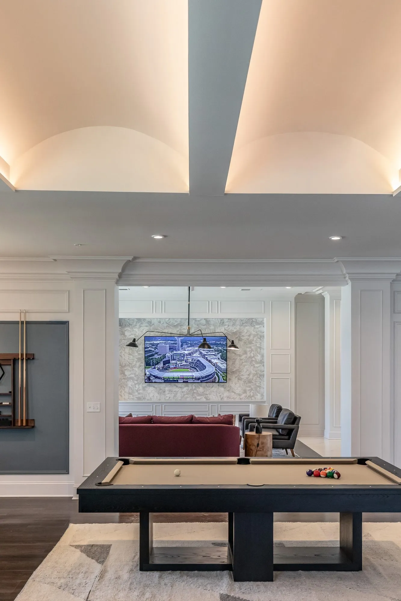 Multifamily amenity clubroom with a billiards table under a vaulted tray ceiling, a burgundy sofa and wall screen beyond