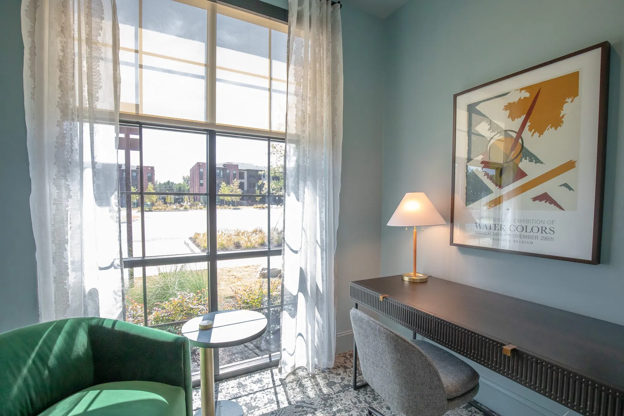 Multifamily interior detail: study desk with a brass lamp and watercolour poster by tall windows, a green velvet armchair