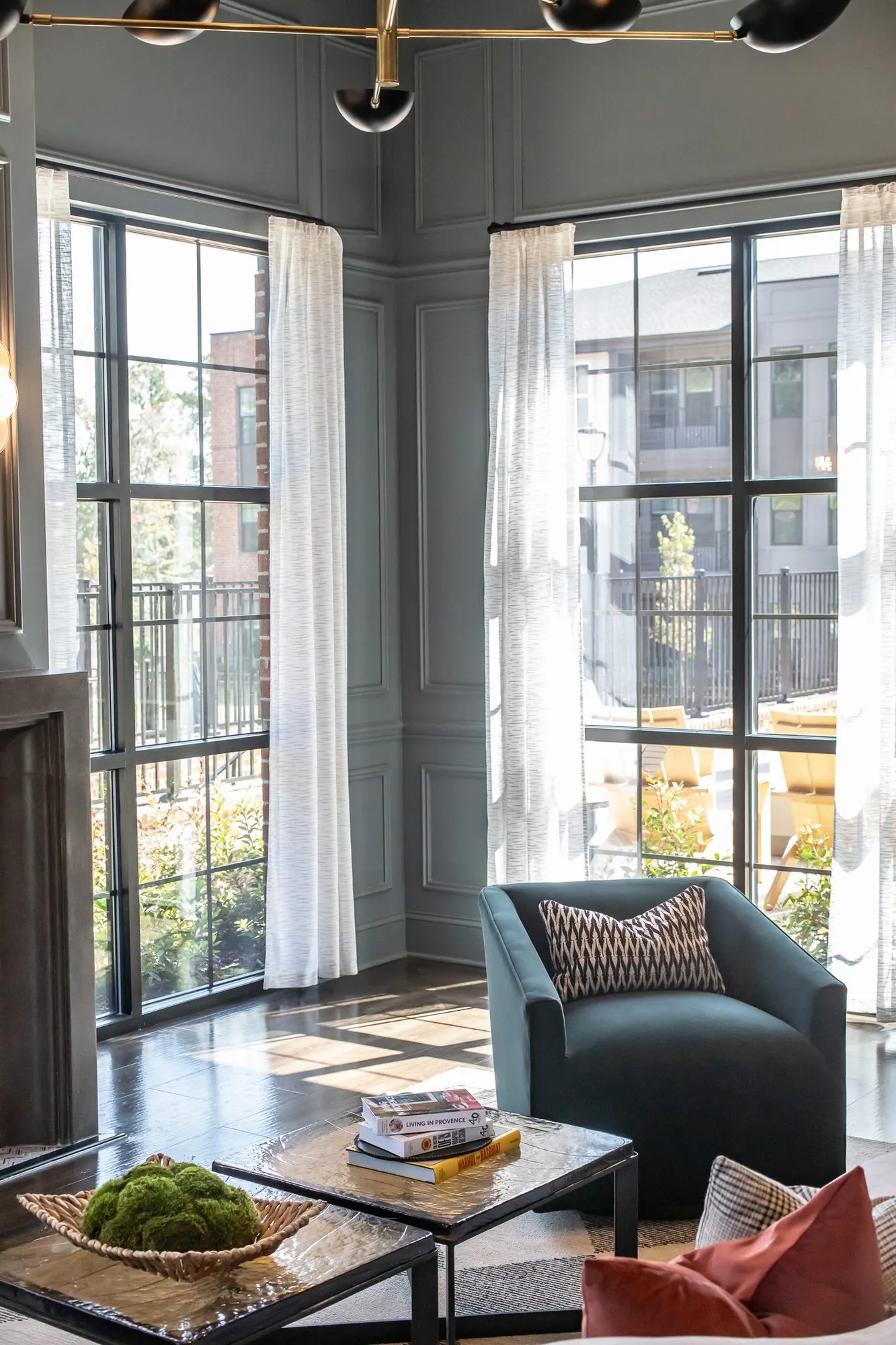 Multifamily amenity lounge with a teal swivel chair by tall black framed windows, a coffee table with a moss bowl