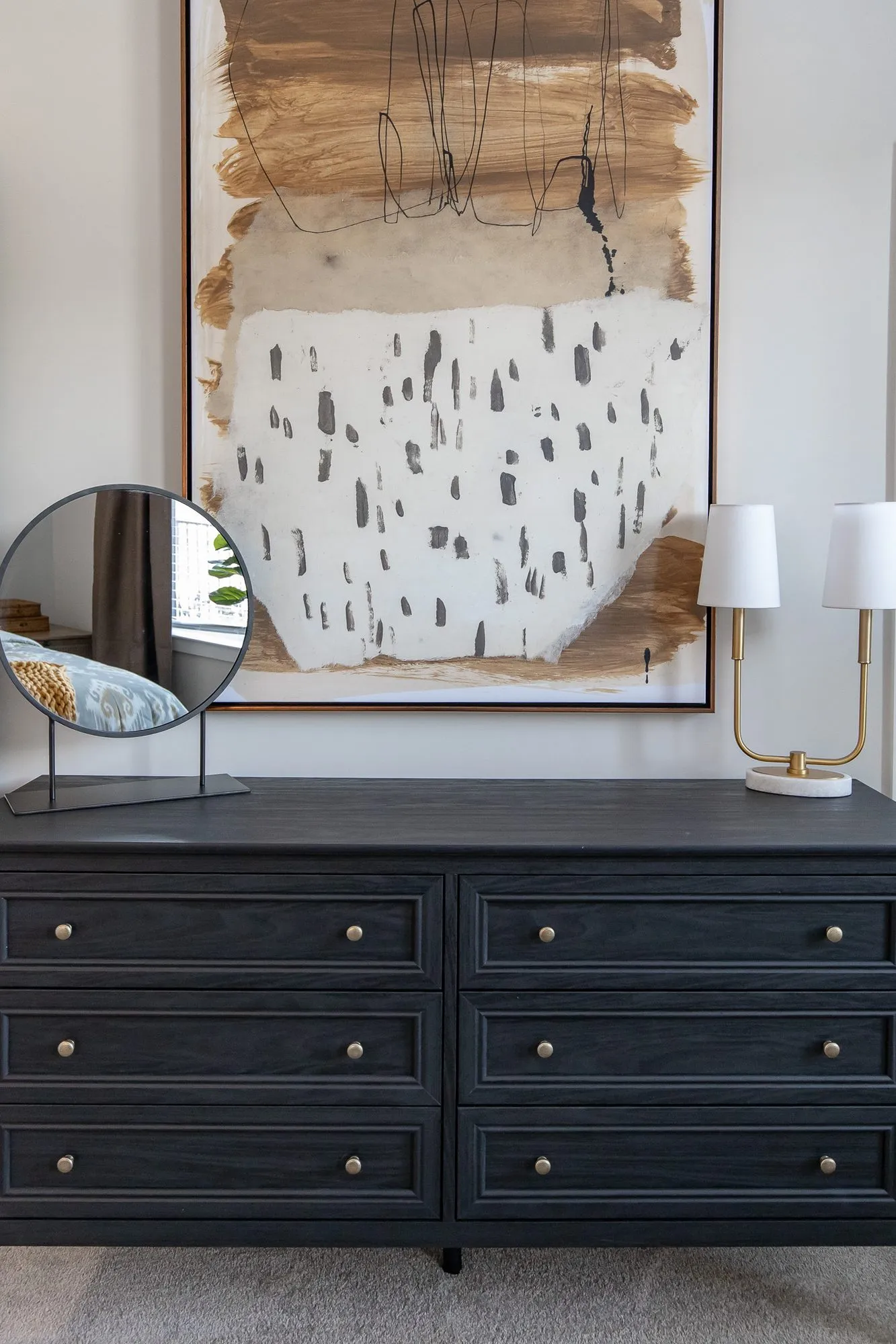 Multifamily interior detail: black dresser below a large ochre and black abstract painting, a round mirror and brass lamp