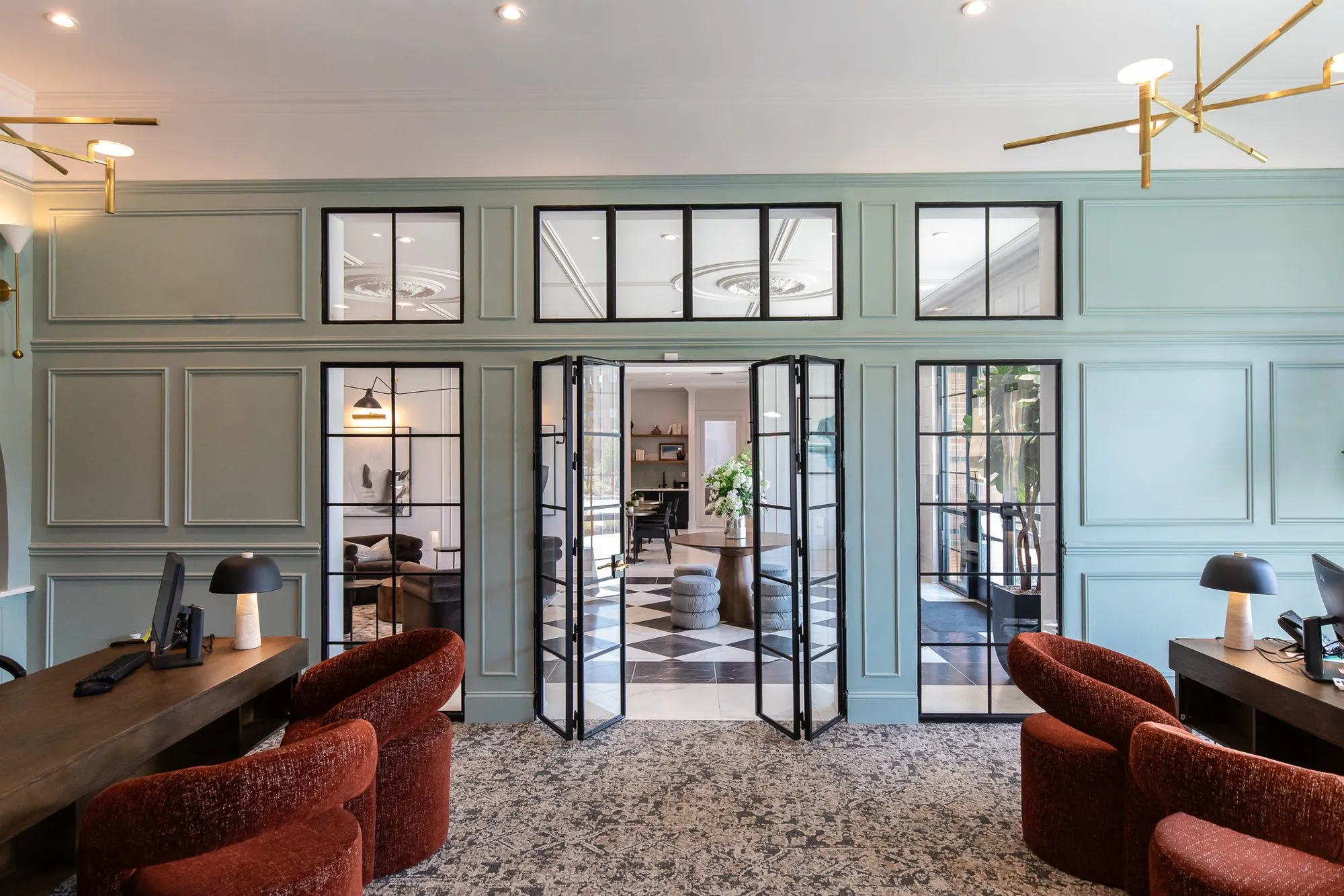 Multifamily amenity leasing office with two desks and rust boucle chairs before sage panelling and black steel French doors