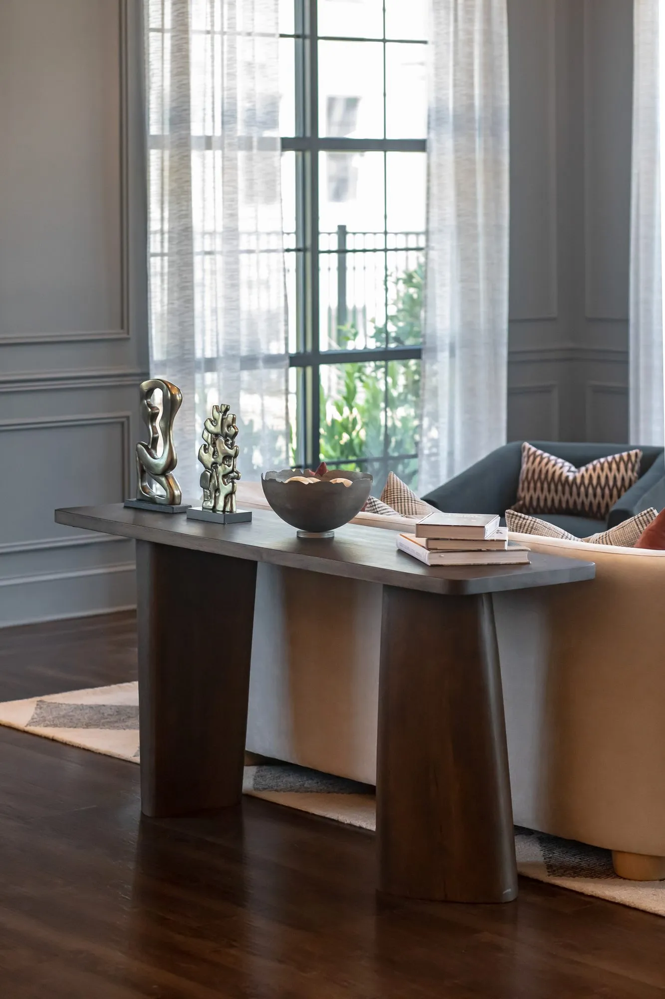 Multifamily amenity sitting room with a walnut console table of sculptures behind a sofa by tall windows and grey panelling