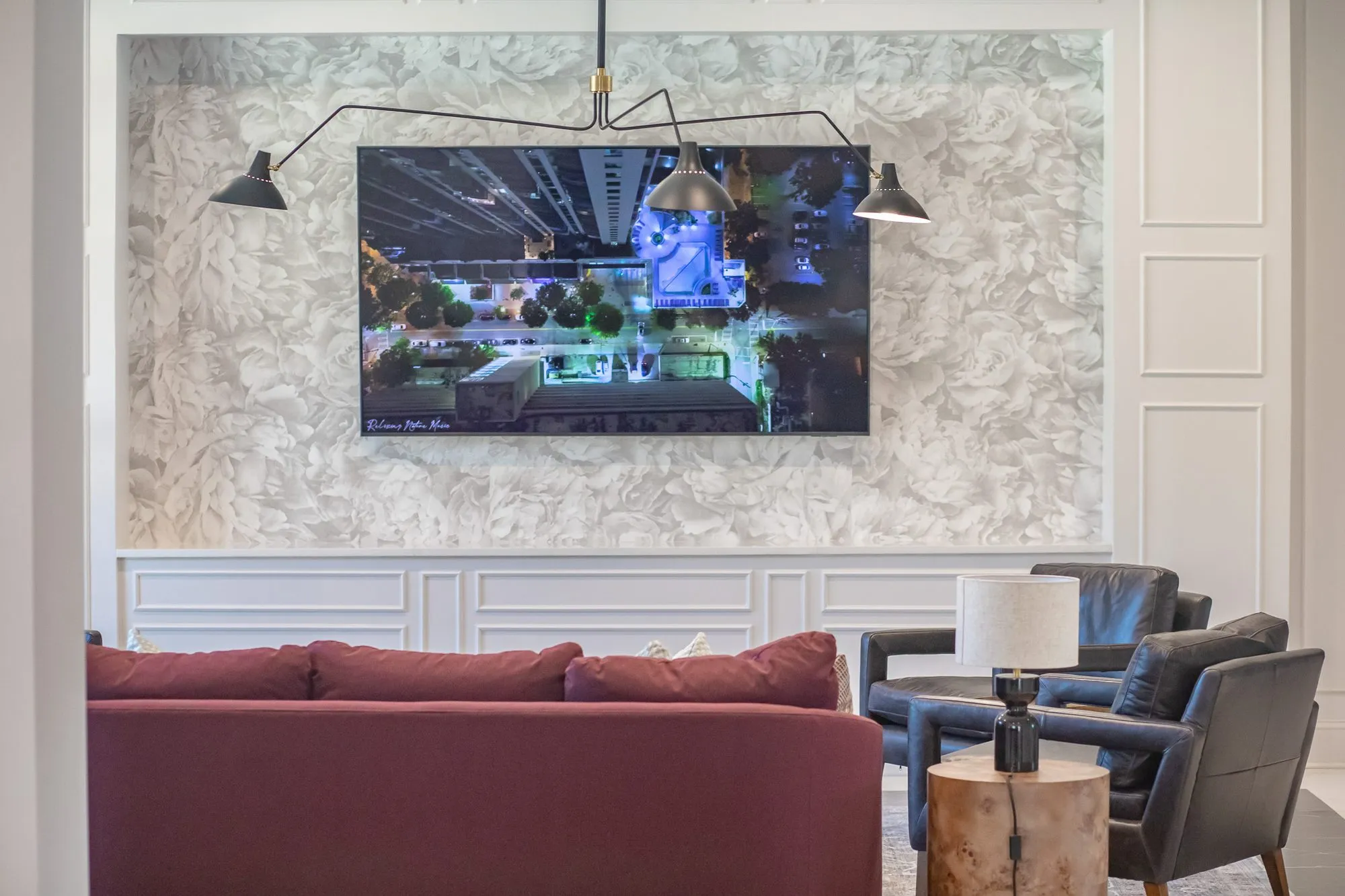 Multifamily amenity clubroom with a wall screen set in white floral relief wallpaper above a burgundy sofa and leather chairs