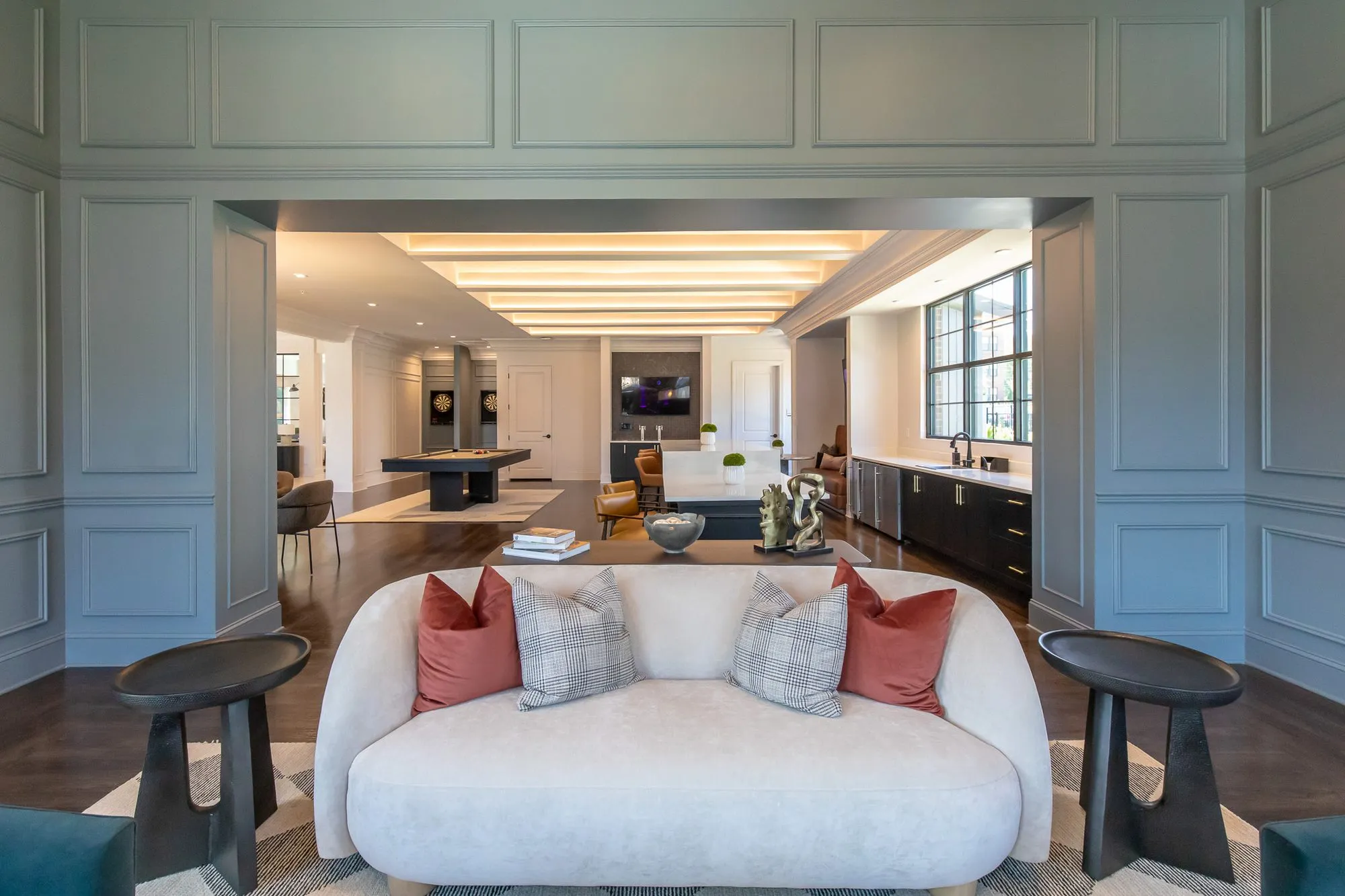 Multifamily amenity sitting room with a white curved sofa and rust and plaid pillows, an opening to the billiards and bar