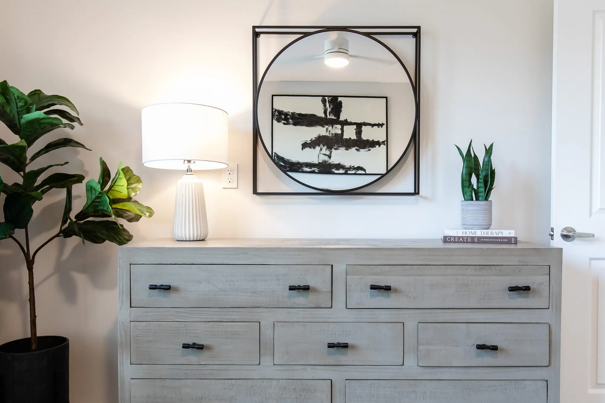 Multifamily interior detail: grey wood dresser with a white lamp below a black circle-framed abstract, a fiddle-leaf fig